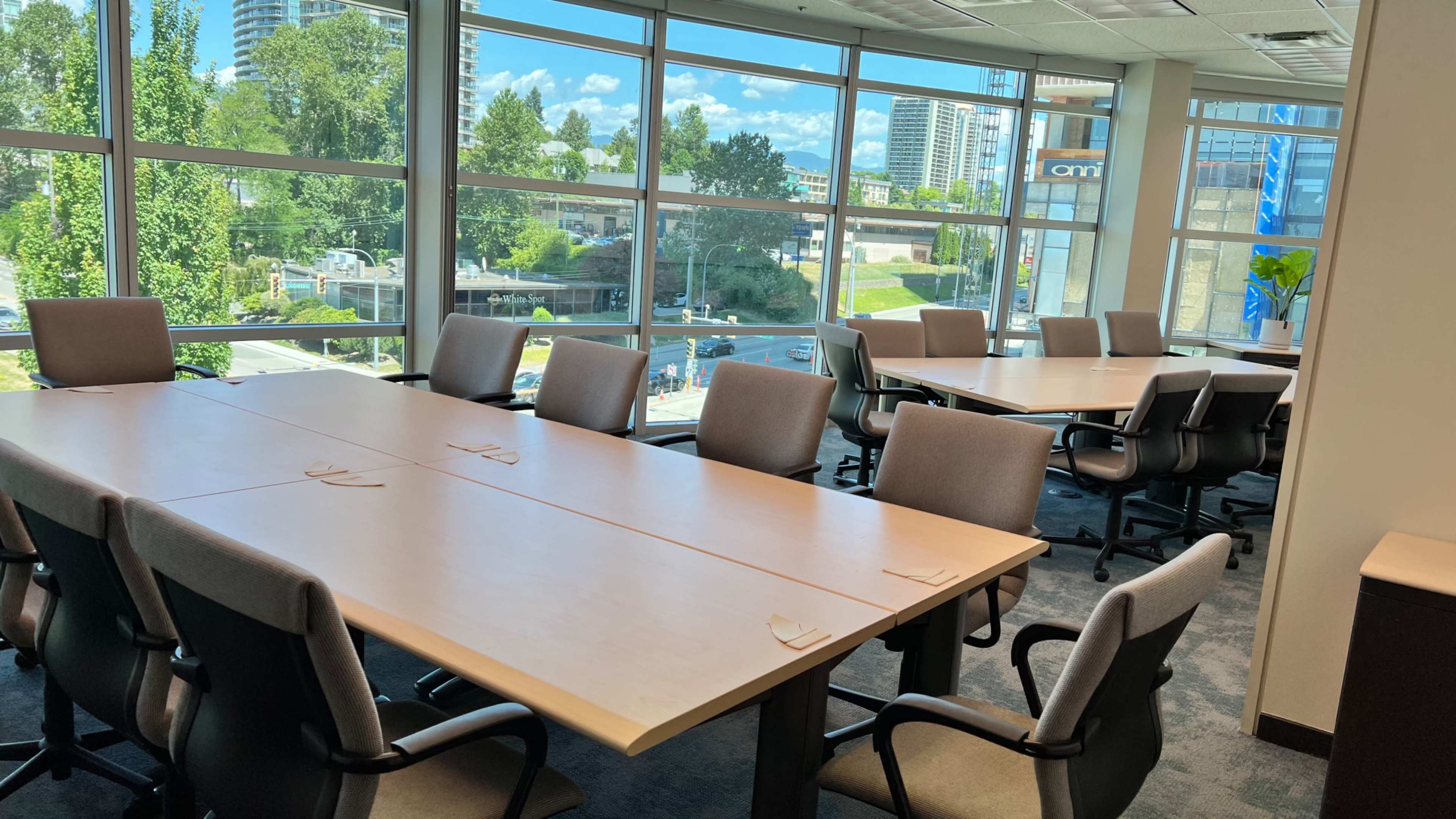 A conference room features two long tables arranged for a meeting, surrounded by chairs, with large windows offering a view of the outdoors.
