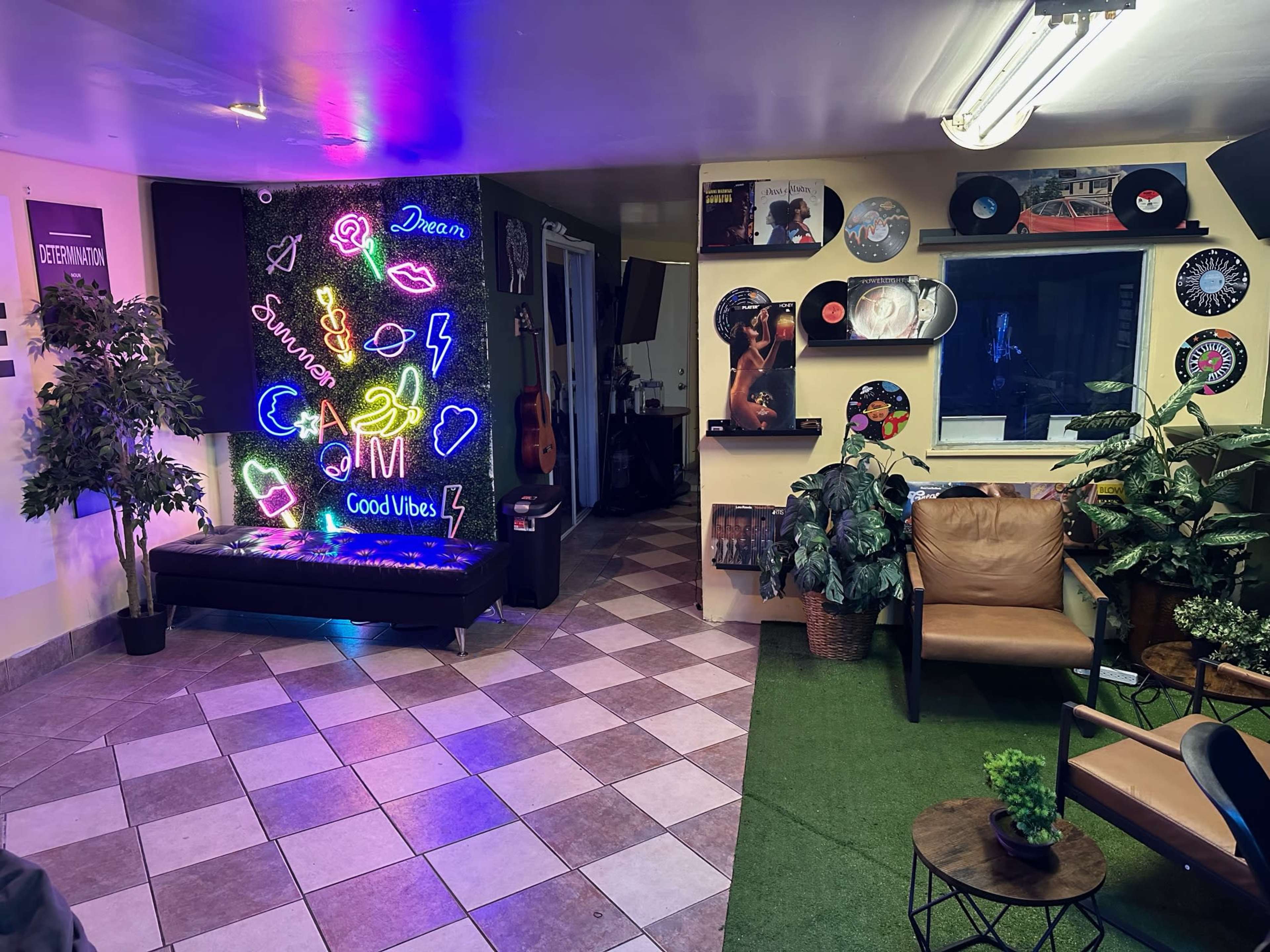 A cozy lounge area with a neon light display, vinyl records on the walls, indoor plants, and comfortable seating arrangements.