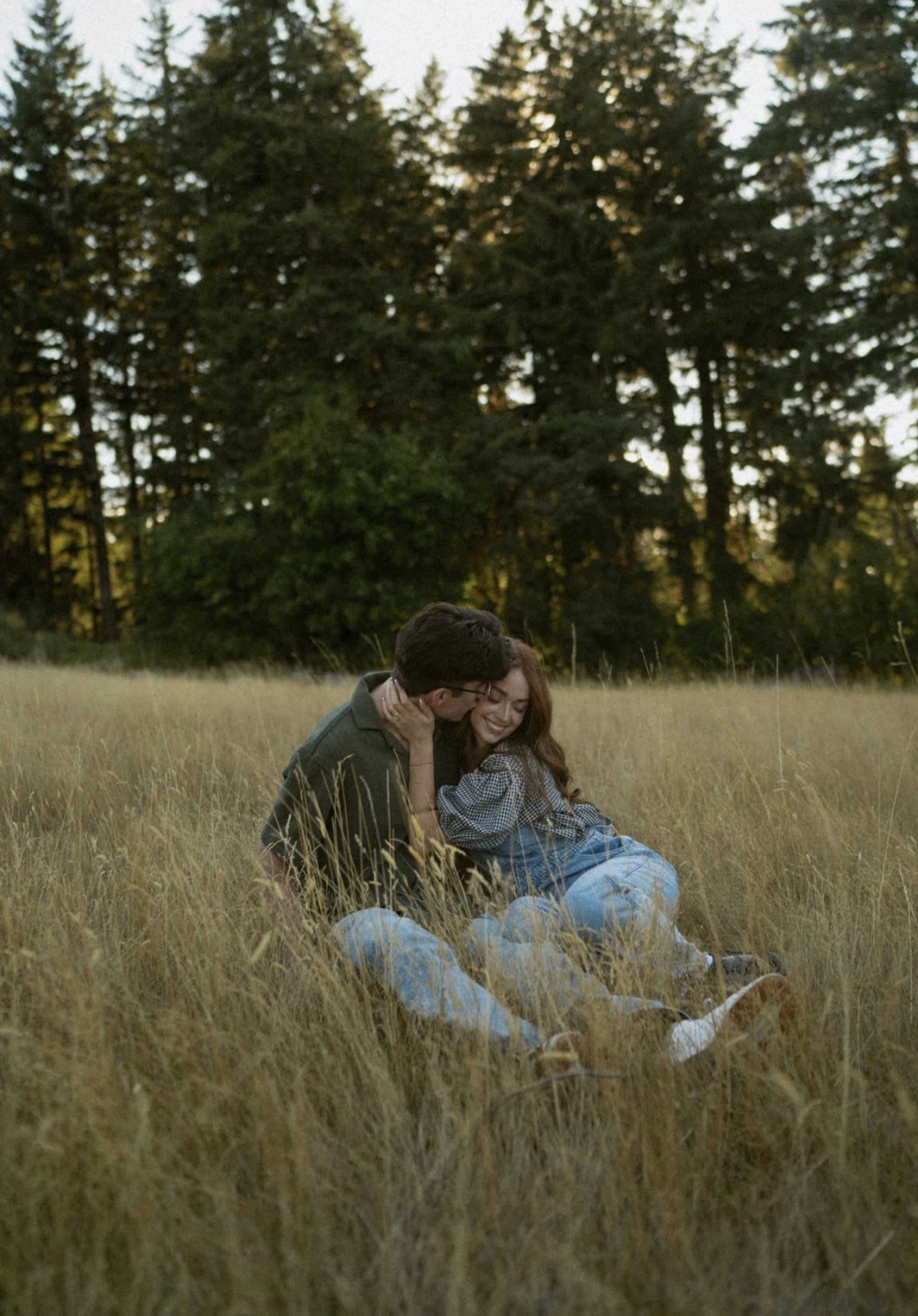 A couple sits in a grassy field surrounded by tall trees, with the sun setting behind them.