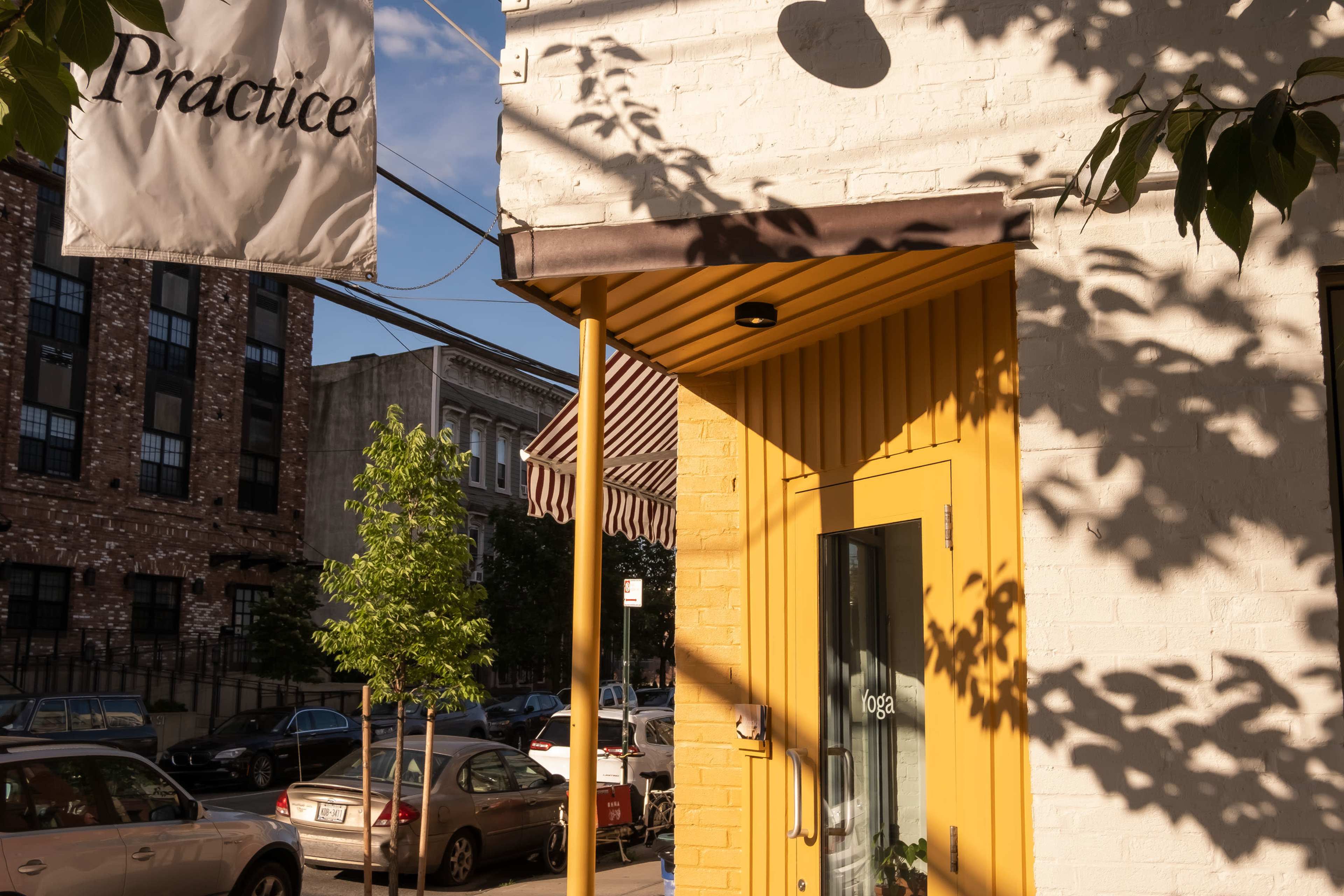 A yellow entrance door to a building with a sign that reads "Practice," accompanied by a striped awning and trees lining the sidewalk.