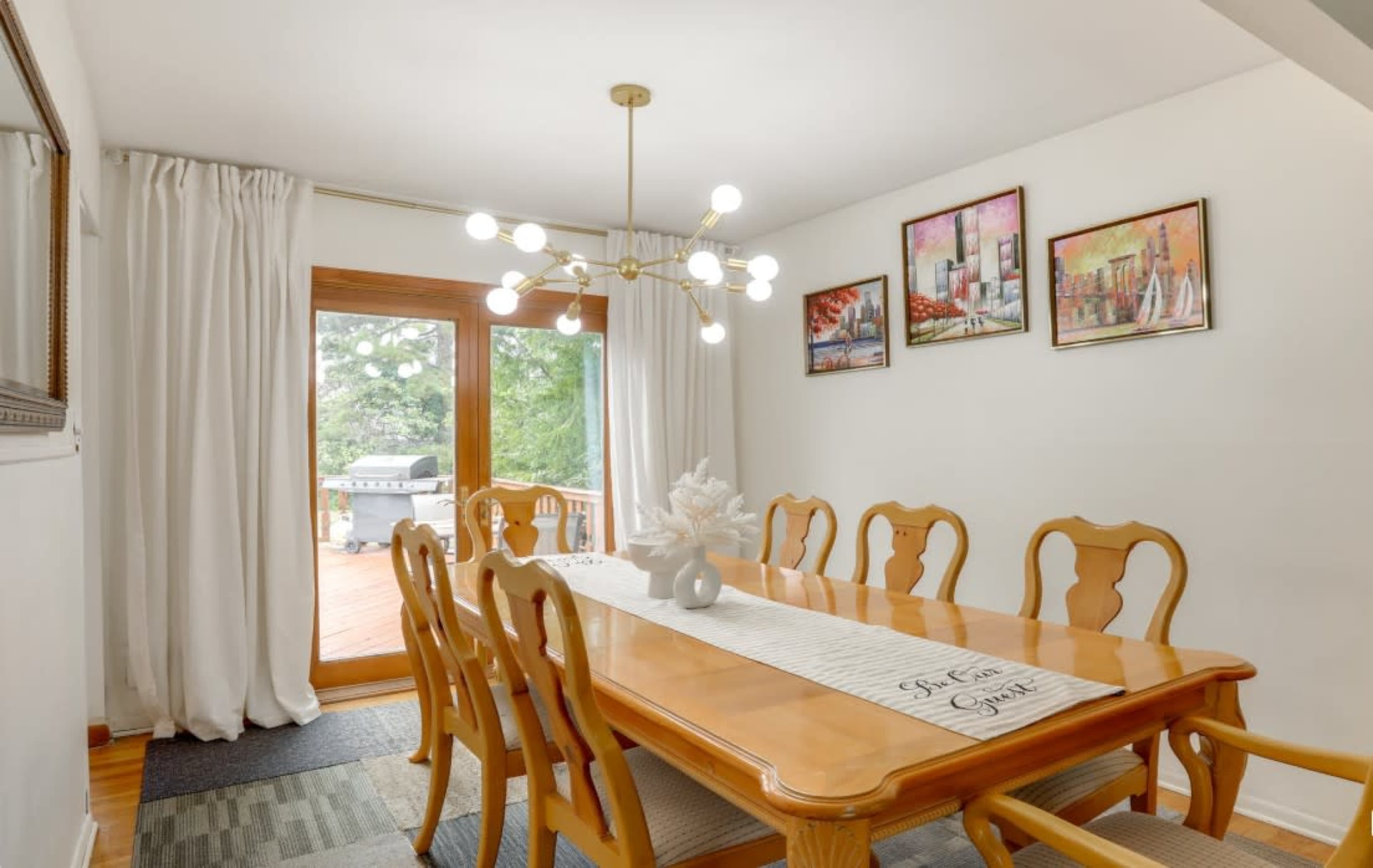 A dining room features a wooden table surrounded by chairs, with a chandelier overhead and artwork on the walls, leading to a deck through sliding glass doors.