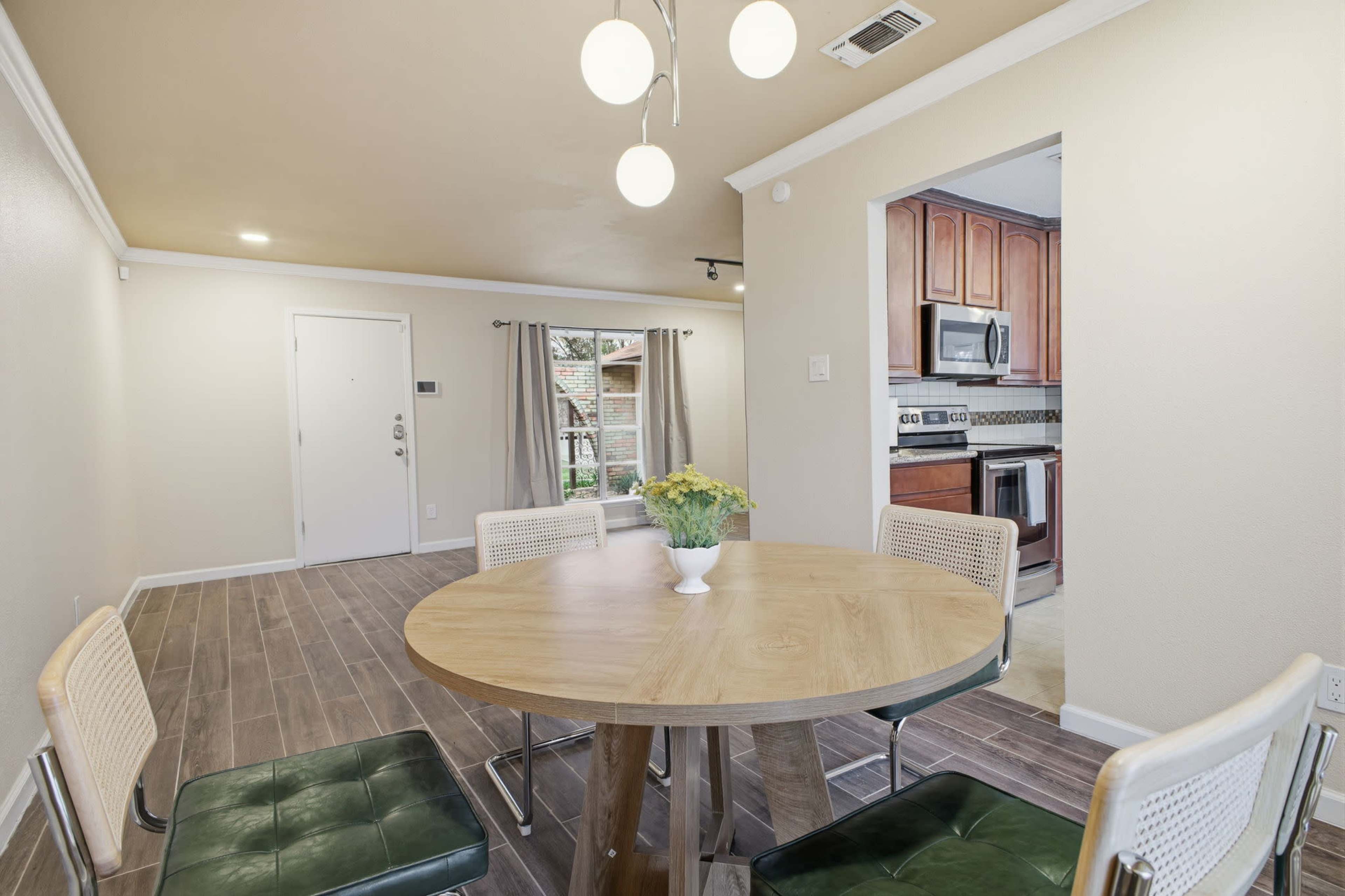 A round dining table with four chairs is set in a well-lit dining area that opens into a kitchen with wooden cabinetry.