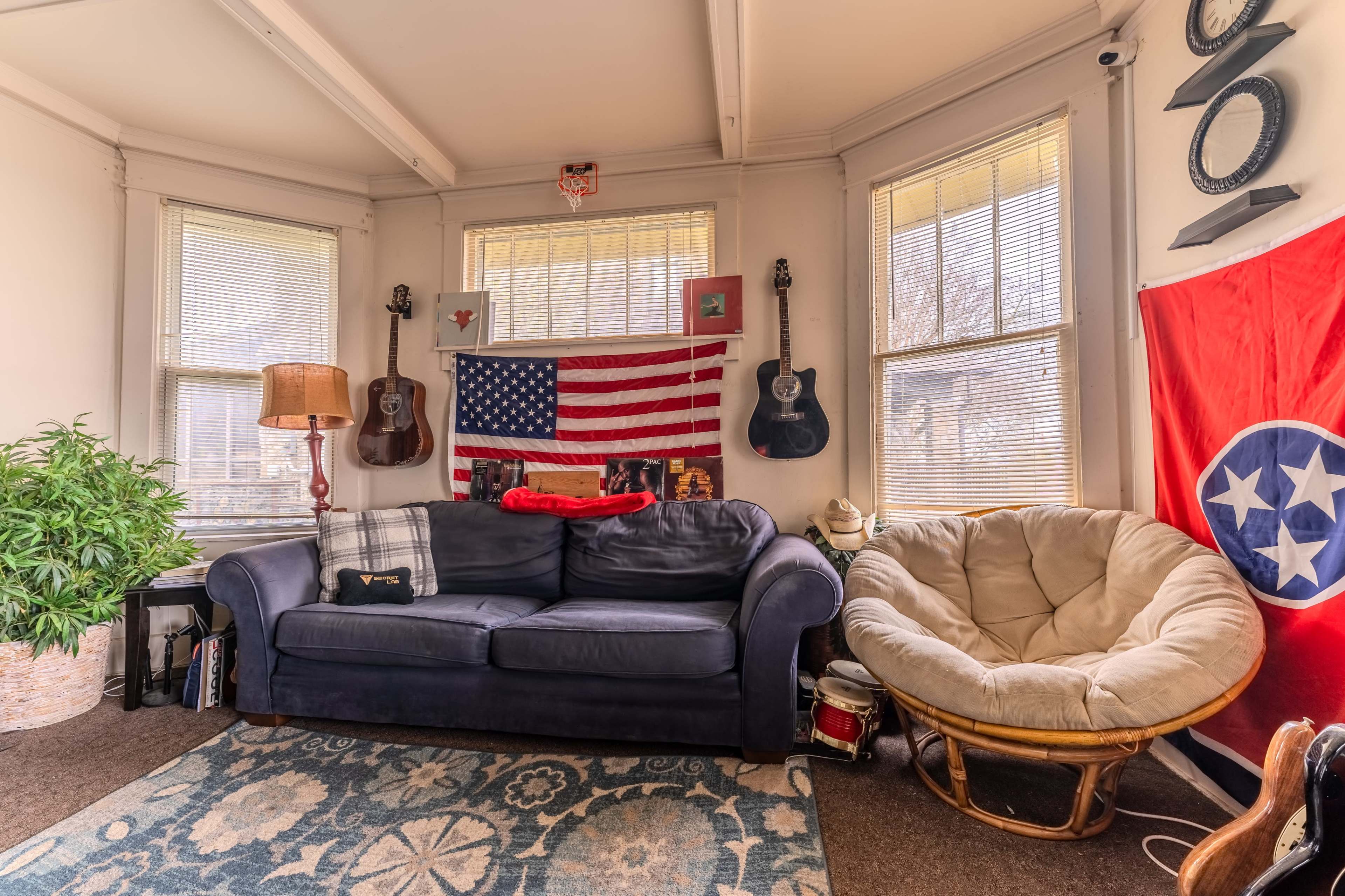 A living room features a dark sofa, a large bean bag chair, an American flag hanging on the wall, and several guitars displayed near the windows.