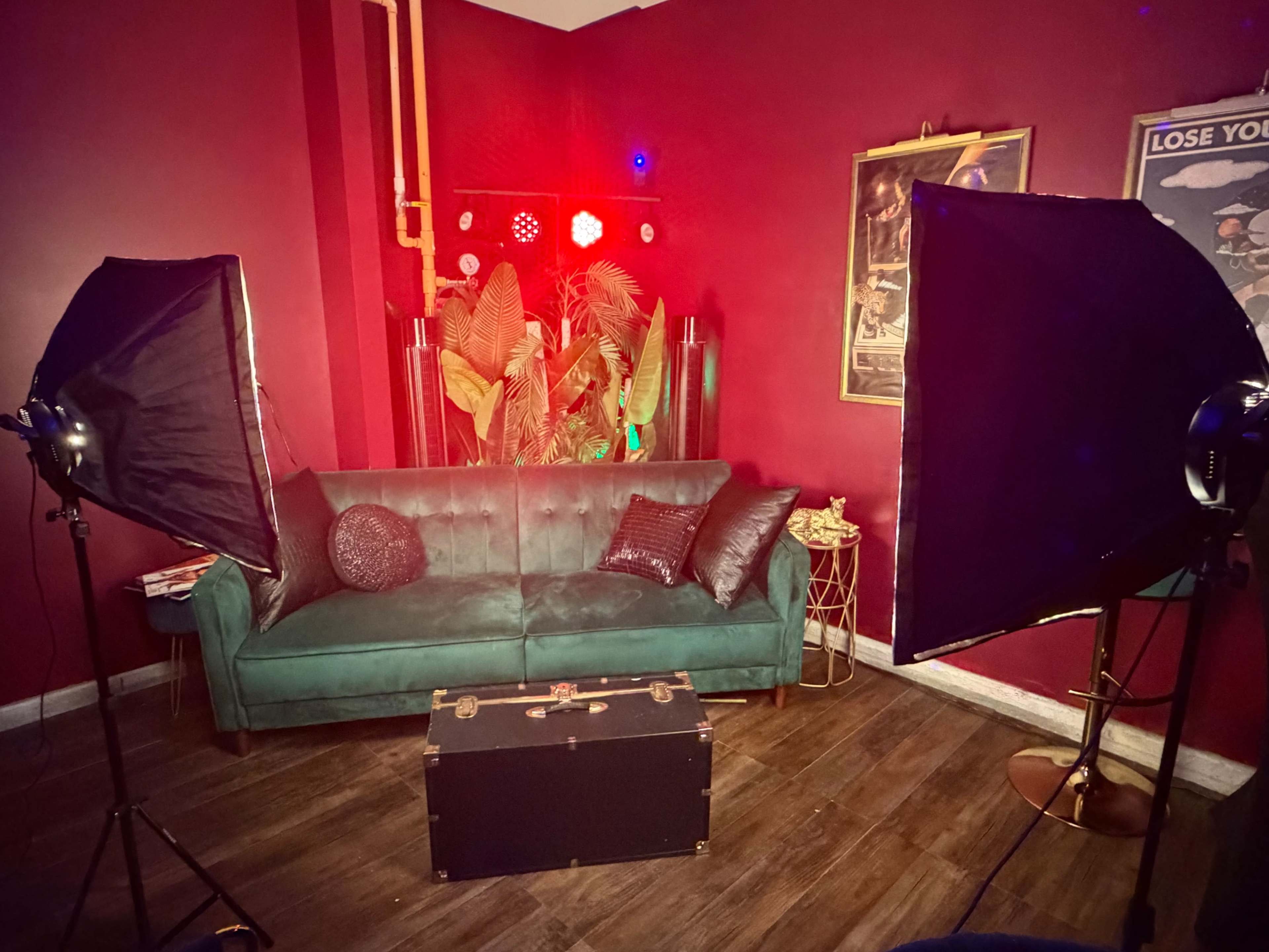 The image shows a cozy, dimly lit room featuring a green sofa, a decorative trunk in front of it, and lighting equipment positioned on either side, with colorful spotlights illuminating the space.