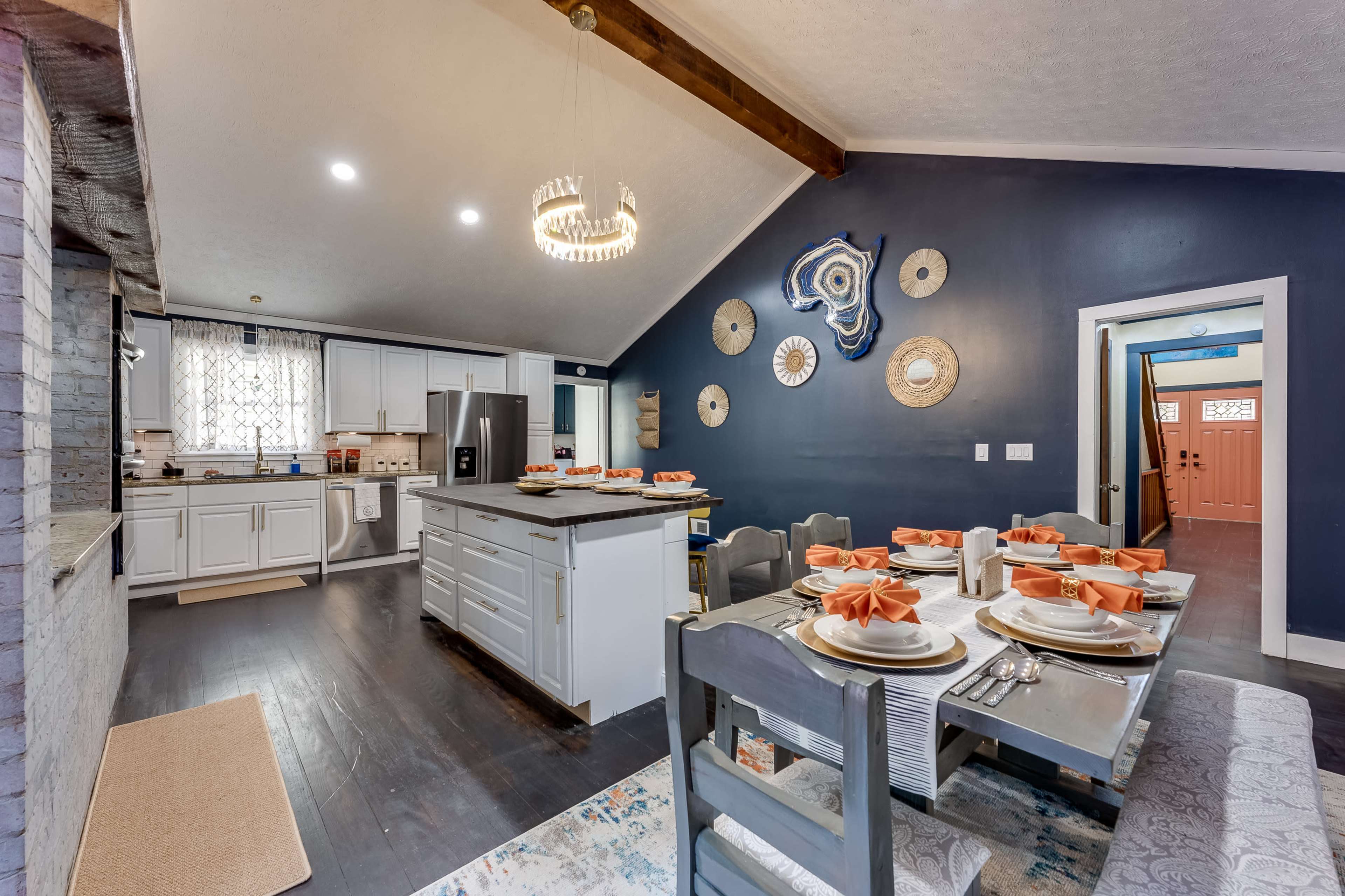 A modern kitchen and dining area with a white table set for six, featuring orange napkins, and a combination of dark blue walls and light cabinetry.