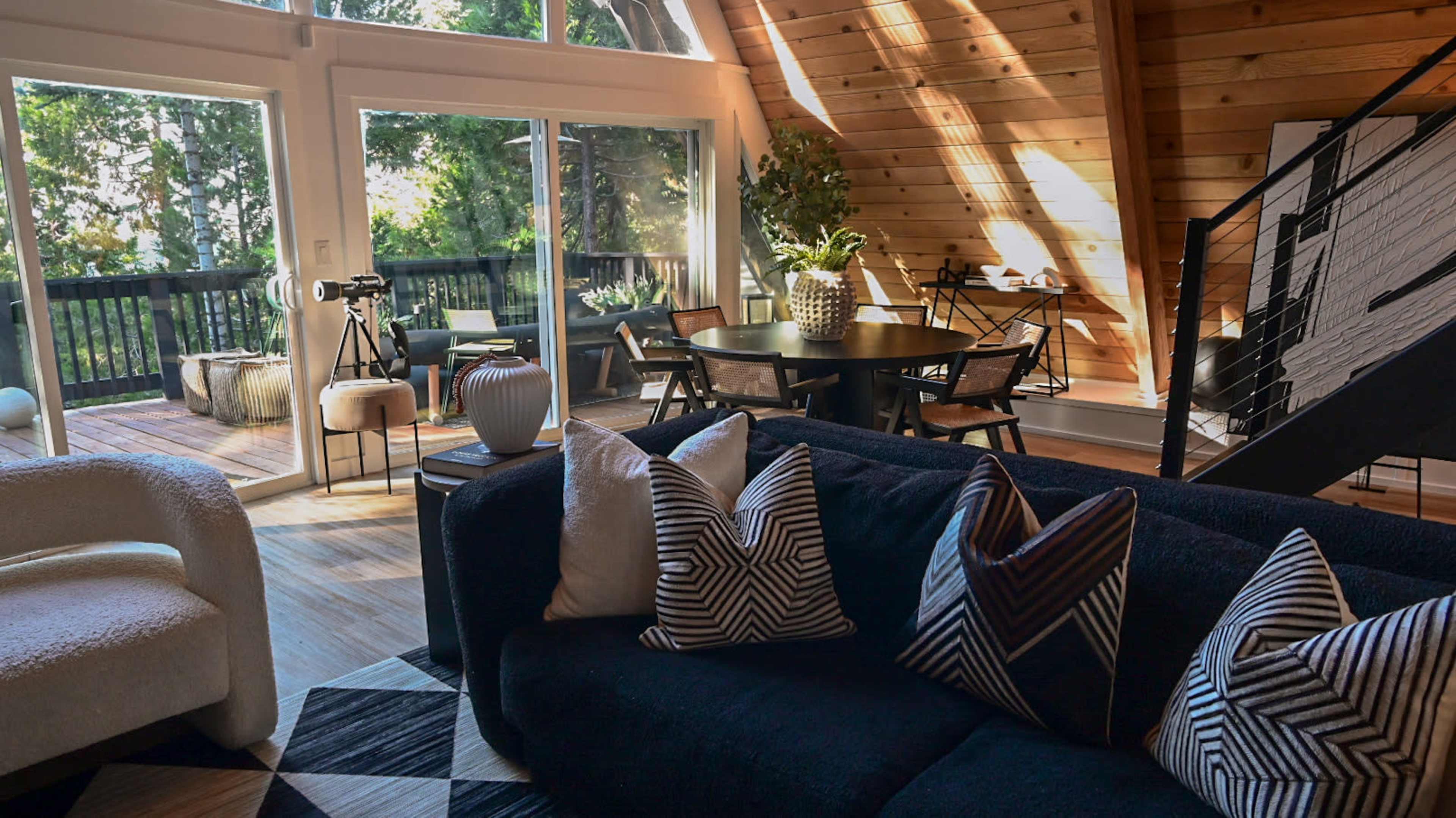 A modern living room with a black sofa featuring patterned cushions, a dining area with a round table, and large windows overlooking a deck surrounded by trees.