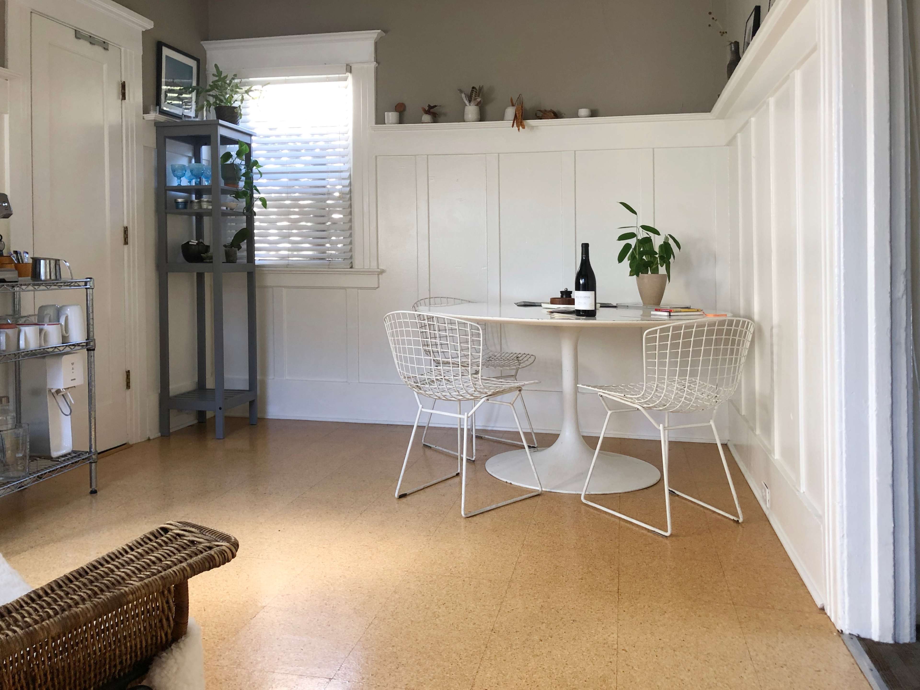 A small dining area features a round white table with two wire chairs, a bottle of wine, and a potted plant, complemented by a shelving unit and light from a window.
