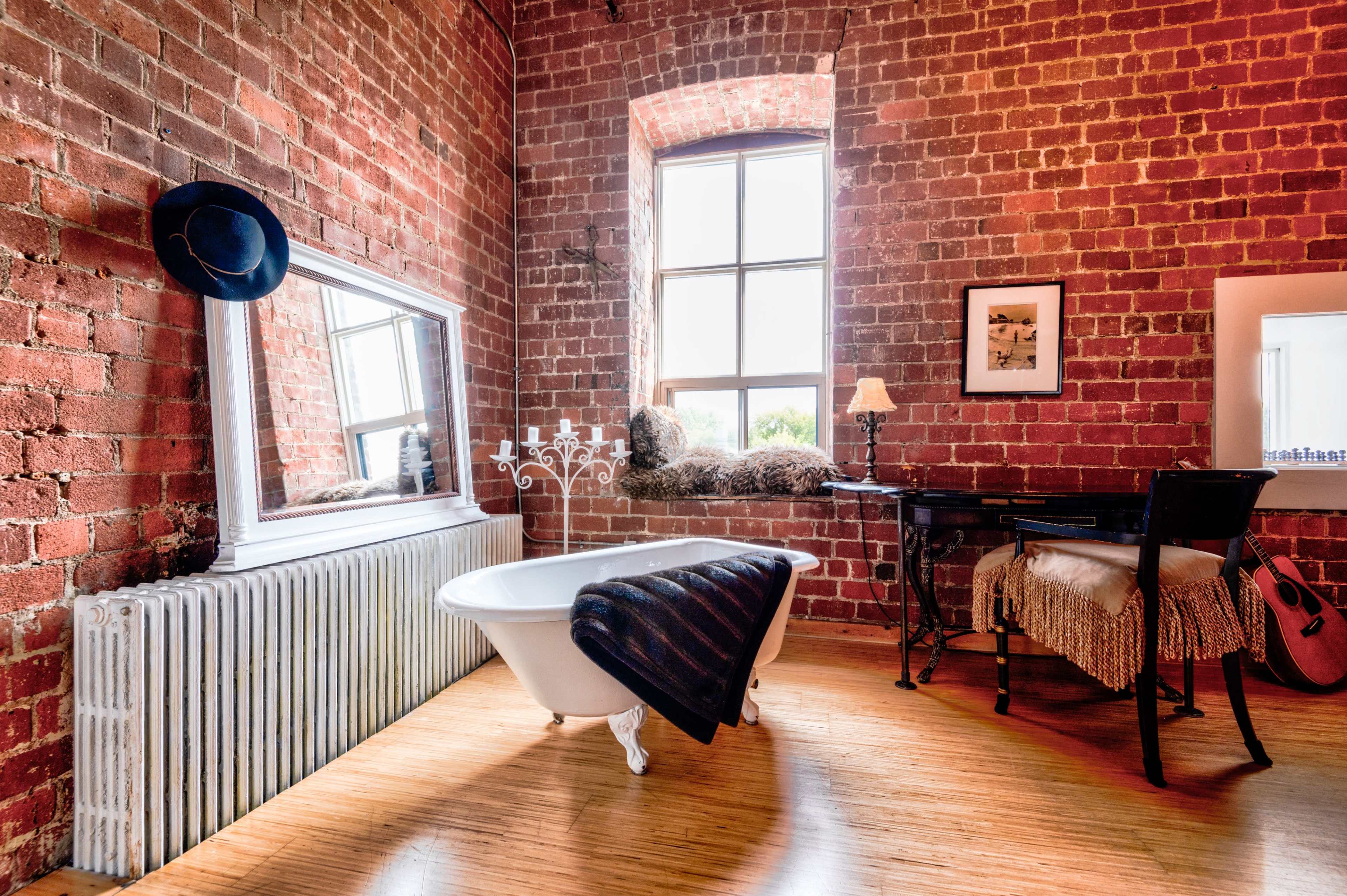 A rustic room featuring a freestanding bathtub, a large mirror, a table with a lamp, and a radiator against exposed brick walls.