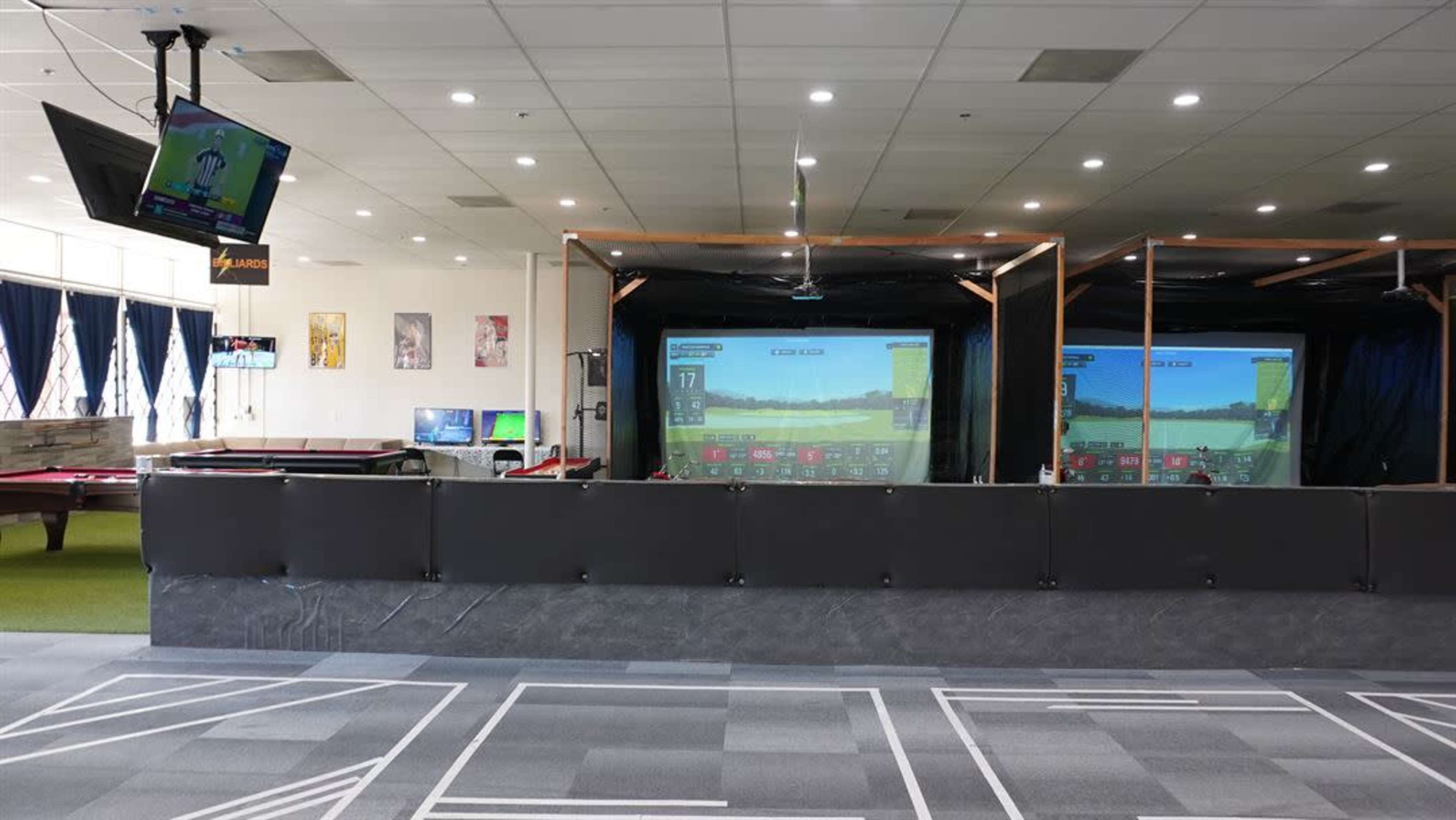 The image shows an indoor golf simulation facility with multiple simulators displaying golf course graphics and a television mounted on the wall.
