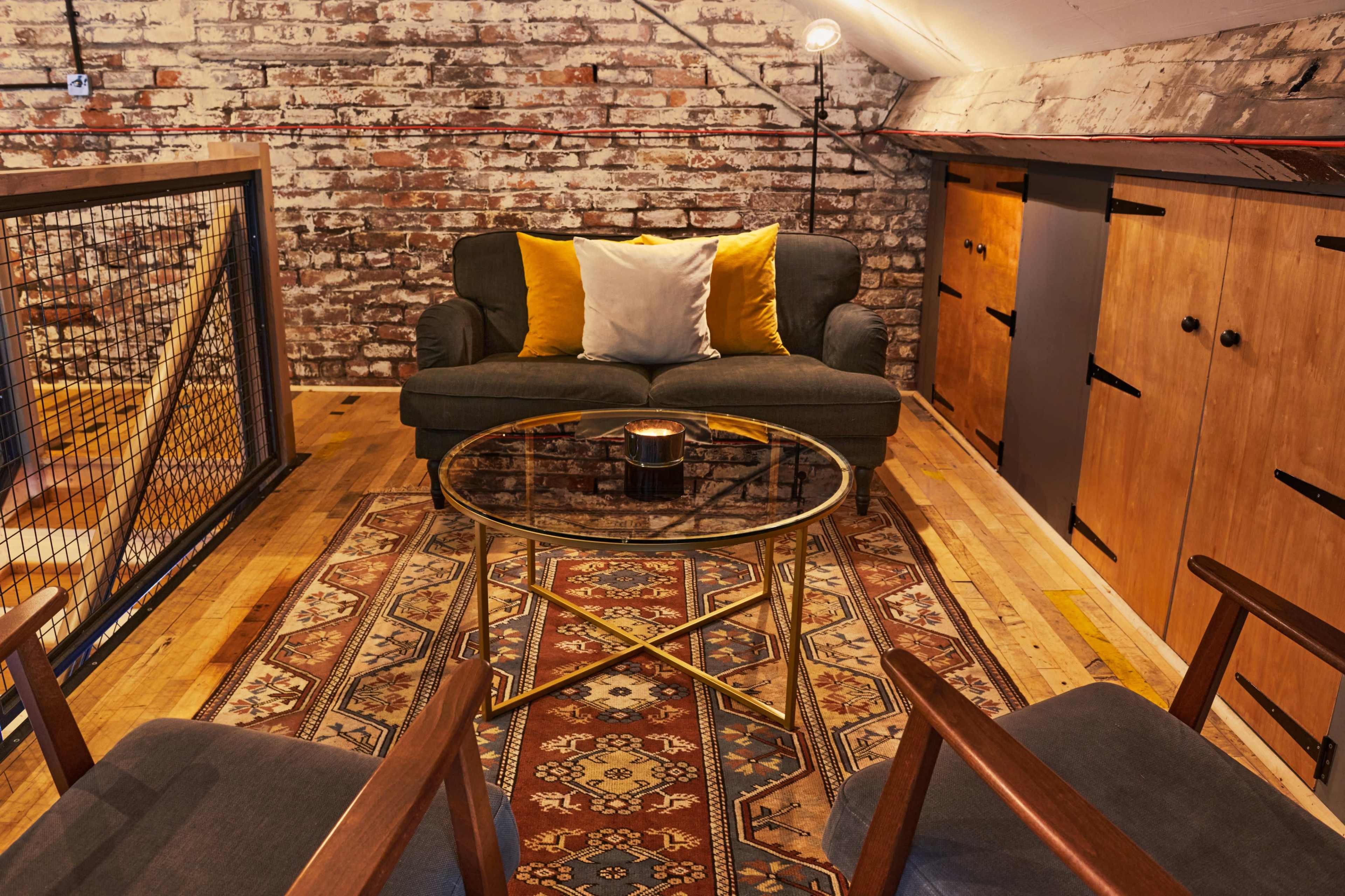 A cozy loft living space with a dark green sofa, two wooden chairs, a round glass coffee table, and exposed brick walls, illuminated by a lamp.