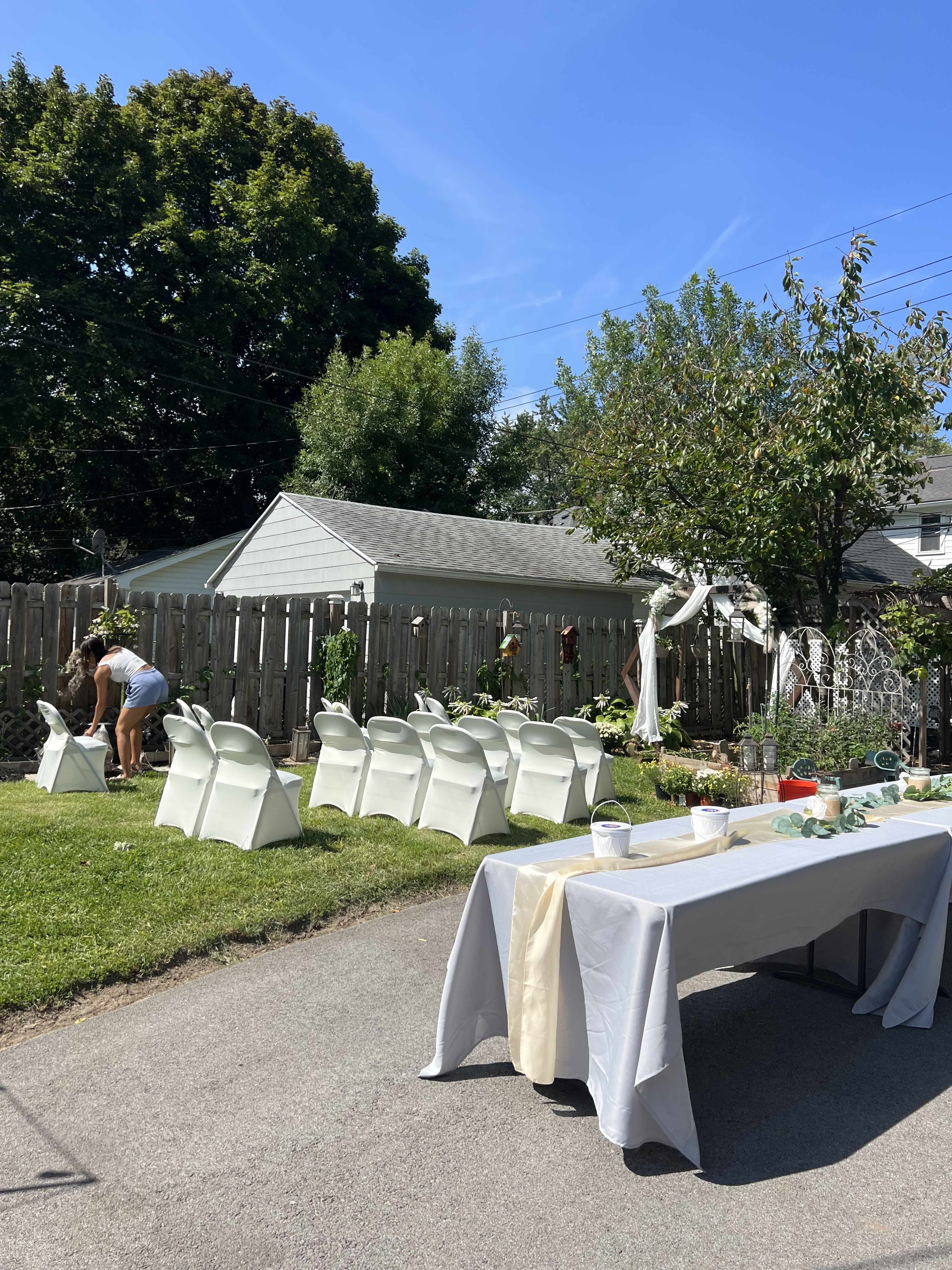 A backyard is set up for an event, featuring white chairs arranged in rows and a table adorned with a beige tablecloth, while a person adjusts decorations nearby.