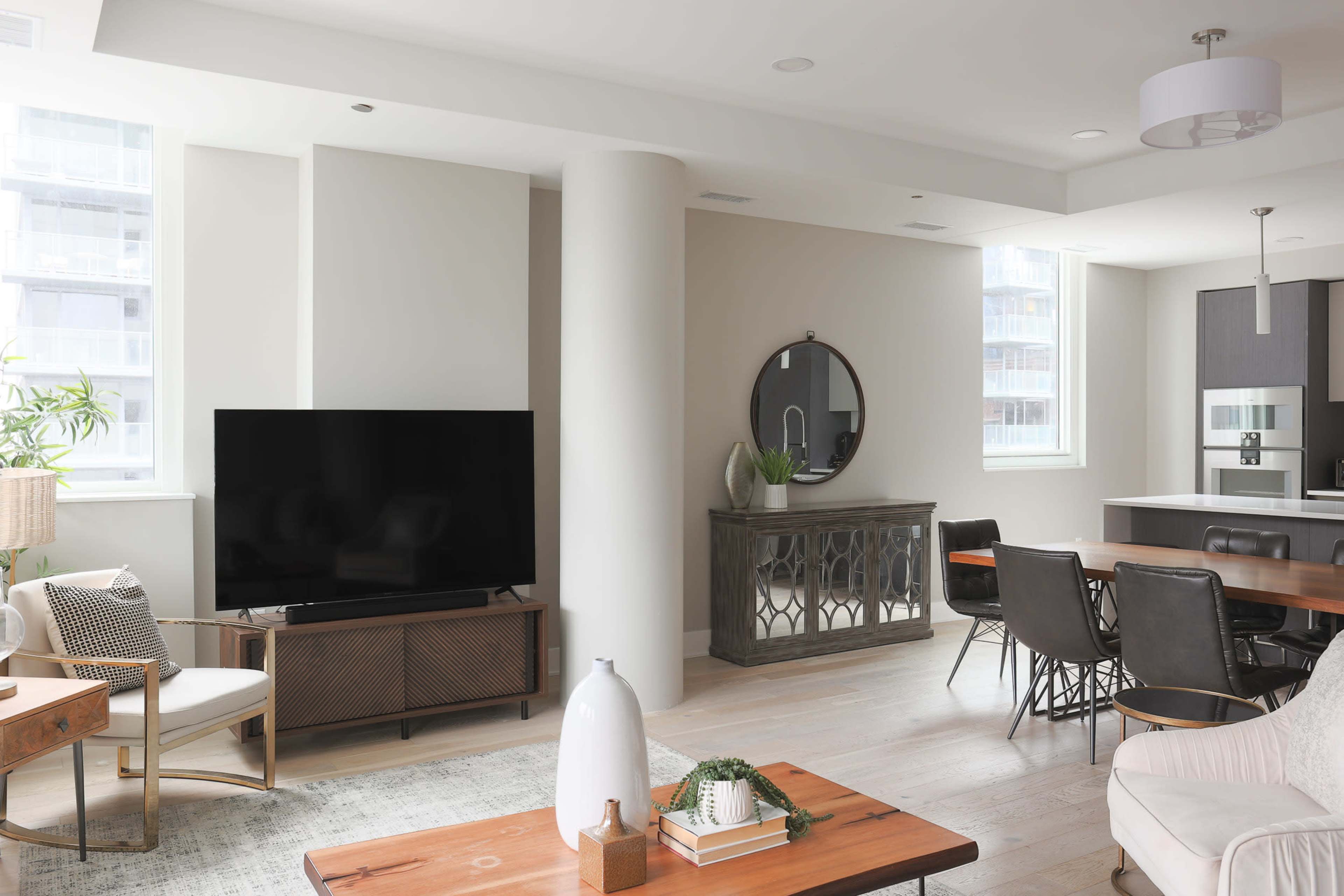 A modern living room featuring a large flat-screen TV, a round mirror on the wall, a wooden dining table, and light-colored furnishings.