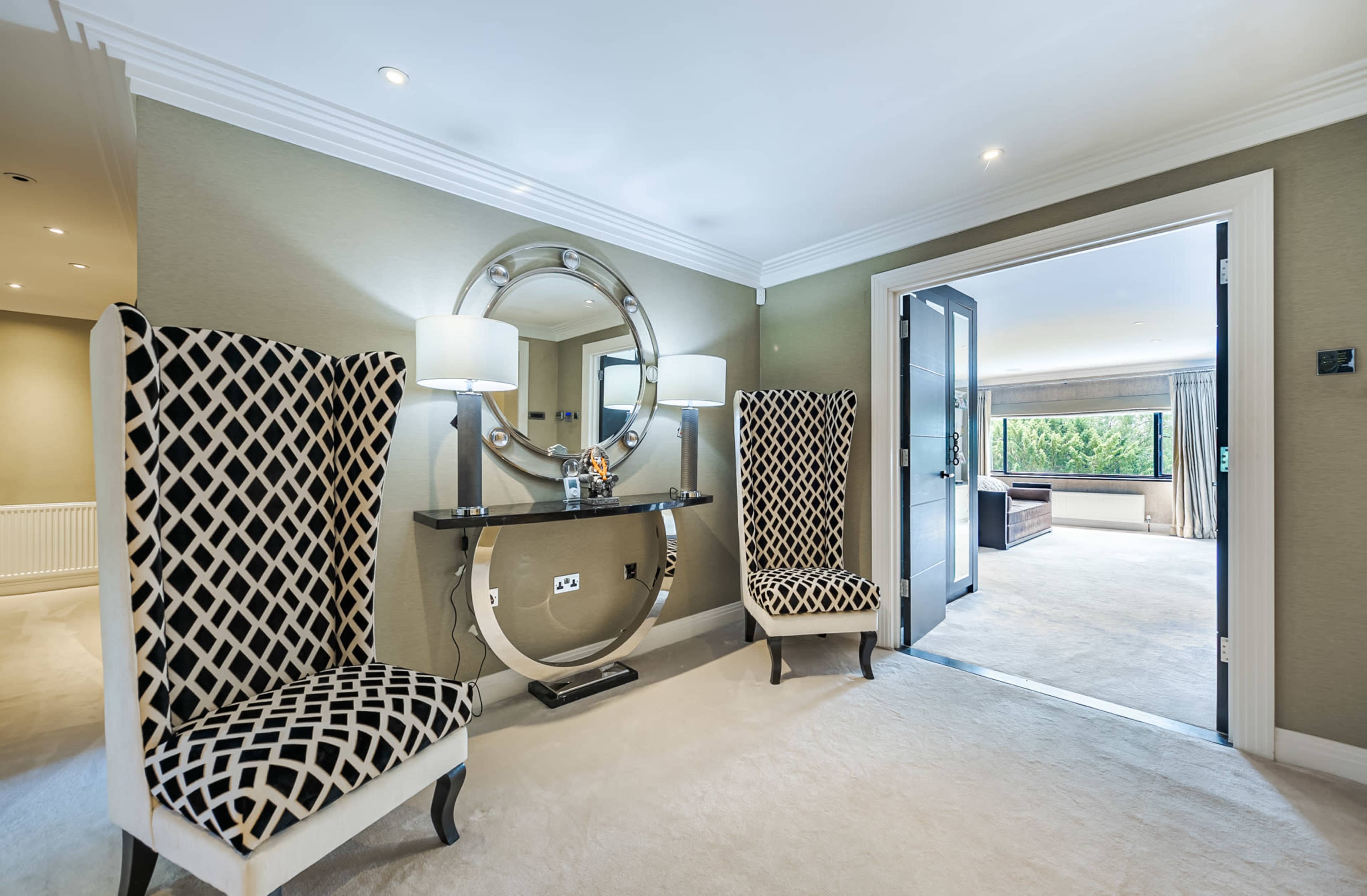 A hallway featuring two patterned chairs flanking a console table, with a round mirror above it and a doorway leading to another room.