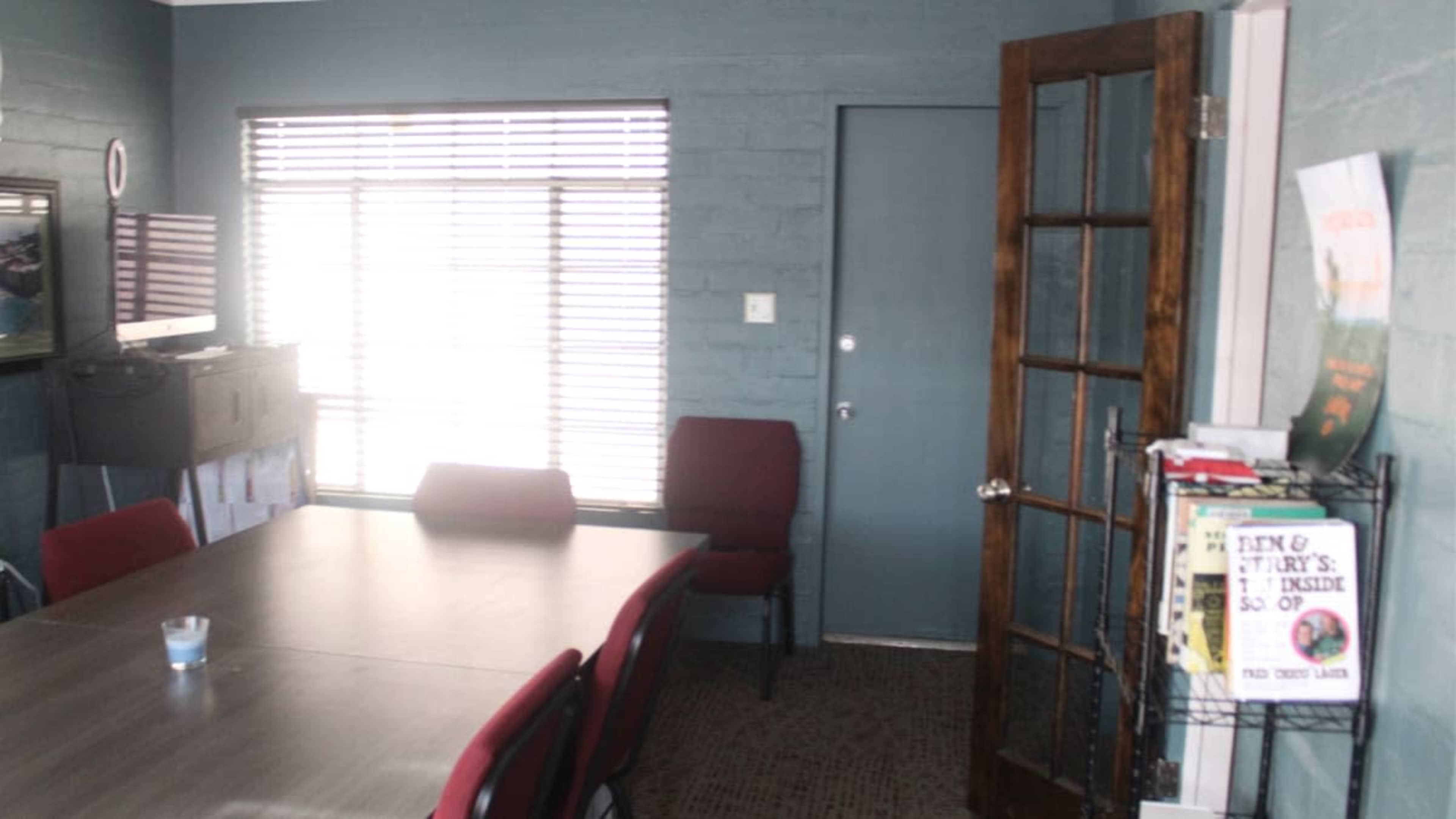 The image shows a small office space with a wooden table surrounded by red chairs, a door leading outside, and a wall organizer filled with papers.
