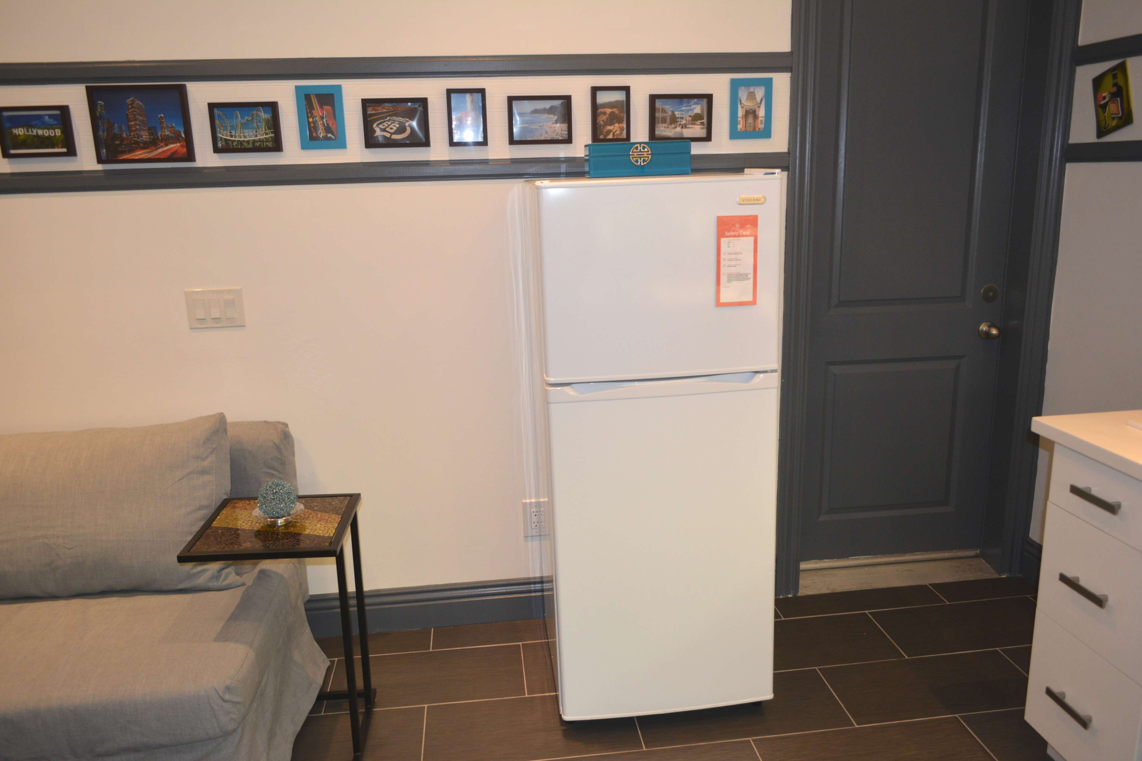 The image shows a white refrigerator positioned next to a couch and framed pictures on the wall above.