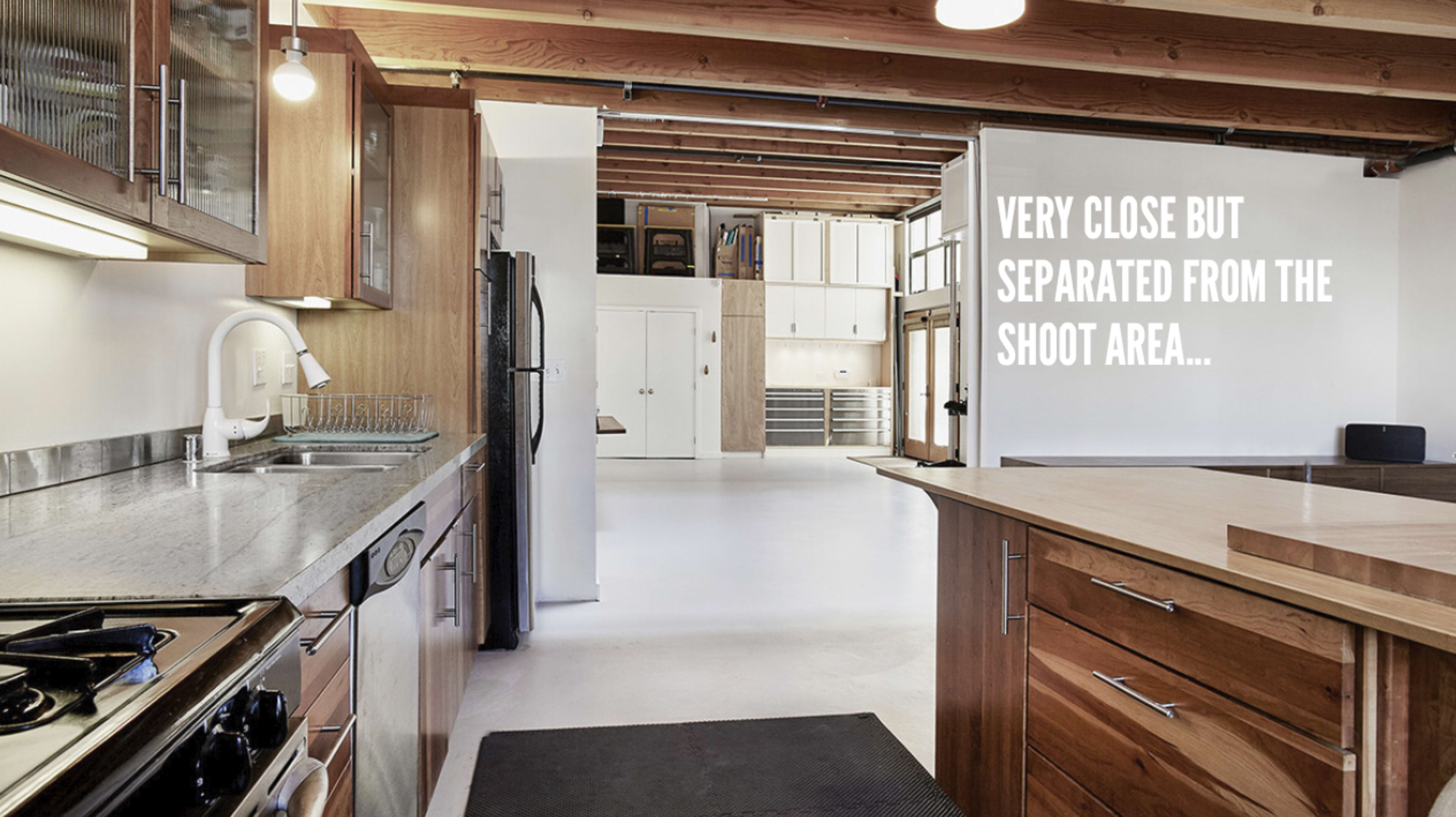 A kitchen with wooden cabinetry and stainless steel appliances, leading into an open, spacious area with white walls and a concrete floor.