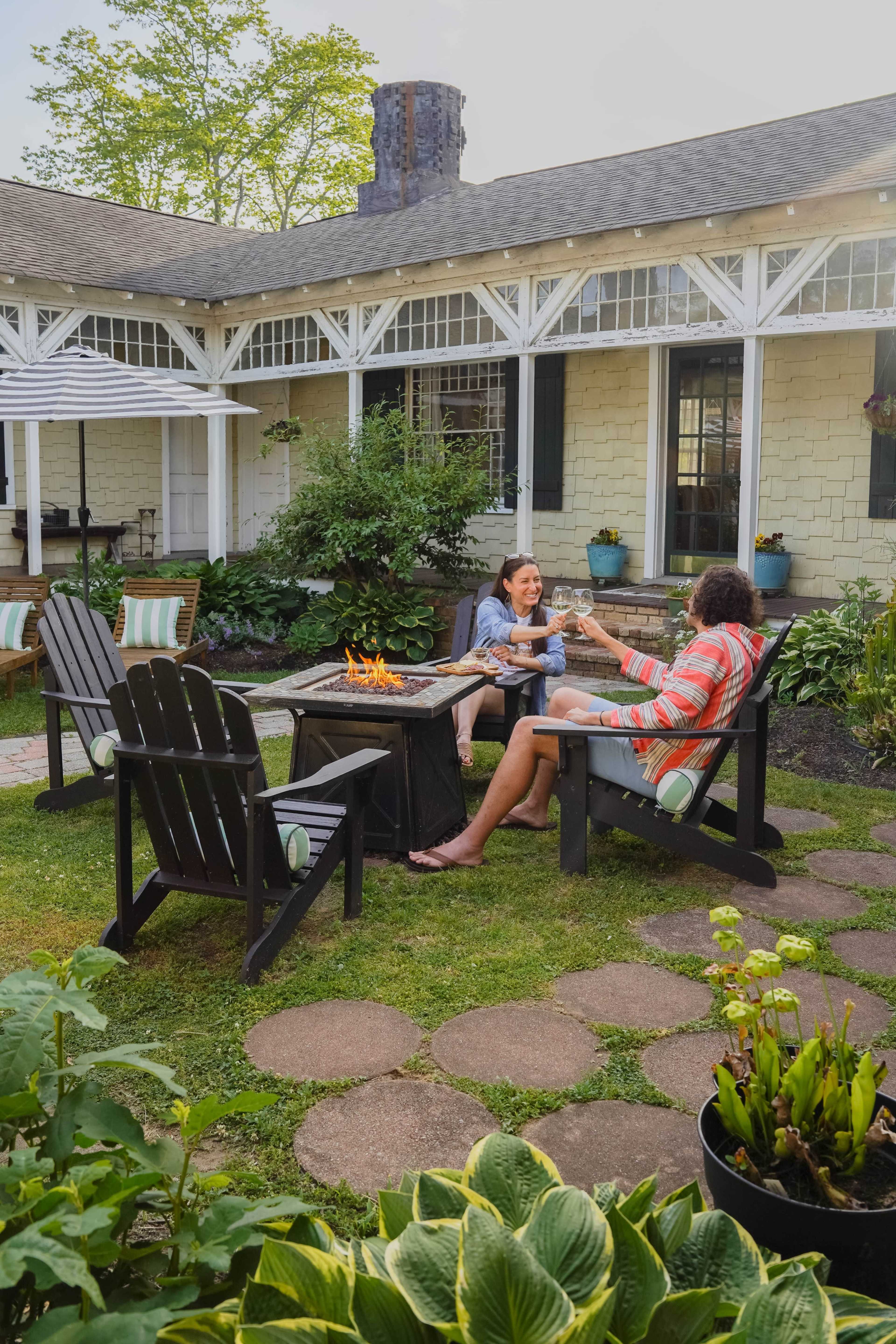 Two people sit by a small fire pit in a garden, toasting drinks while surrounded by greenery and a cozy patio setting.