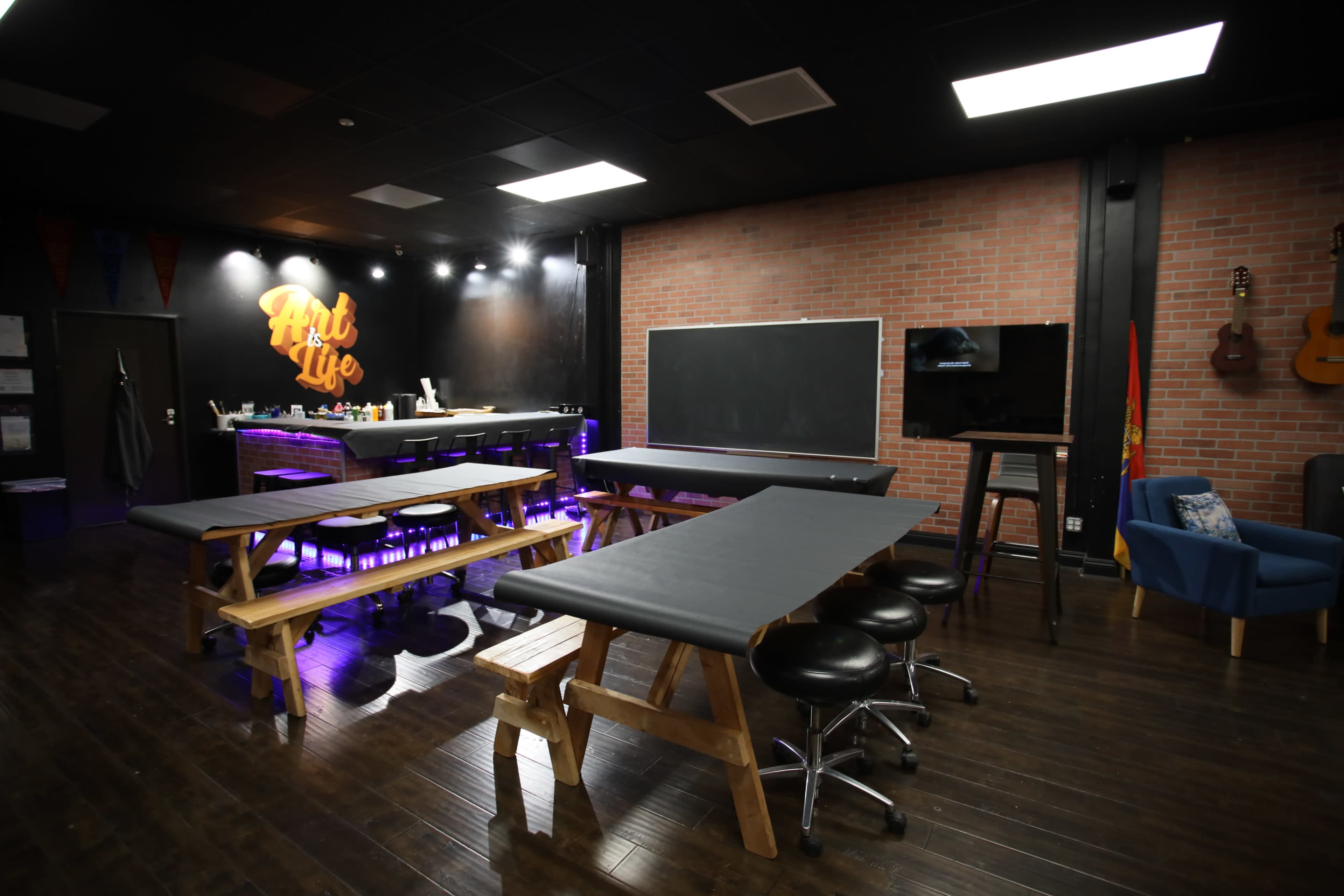 A spacious room designed for art activities, featuring several wooden tables, a blackboard, a television, and a bar area with purple lighting against a brick wall.