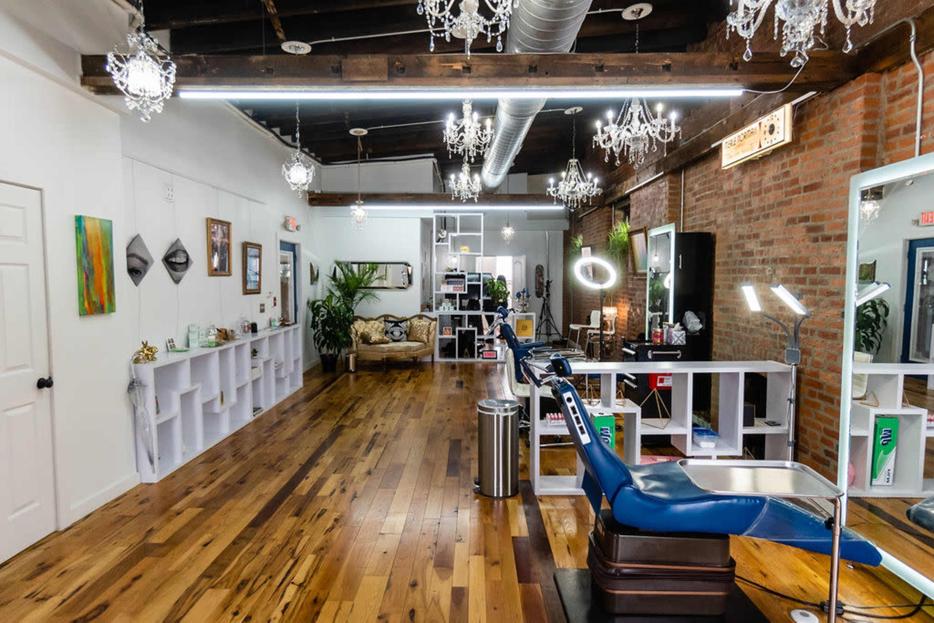 A modern tattoo studio with wooden floors, decorative chandeliers, and a blue tattoo chair in the foreground.