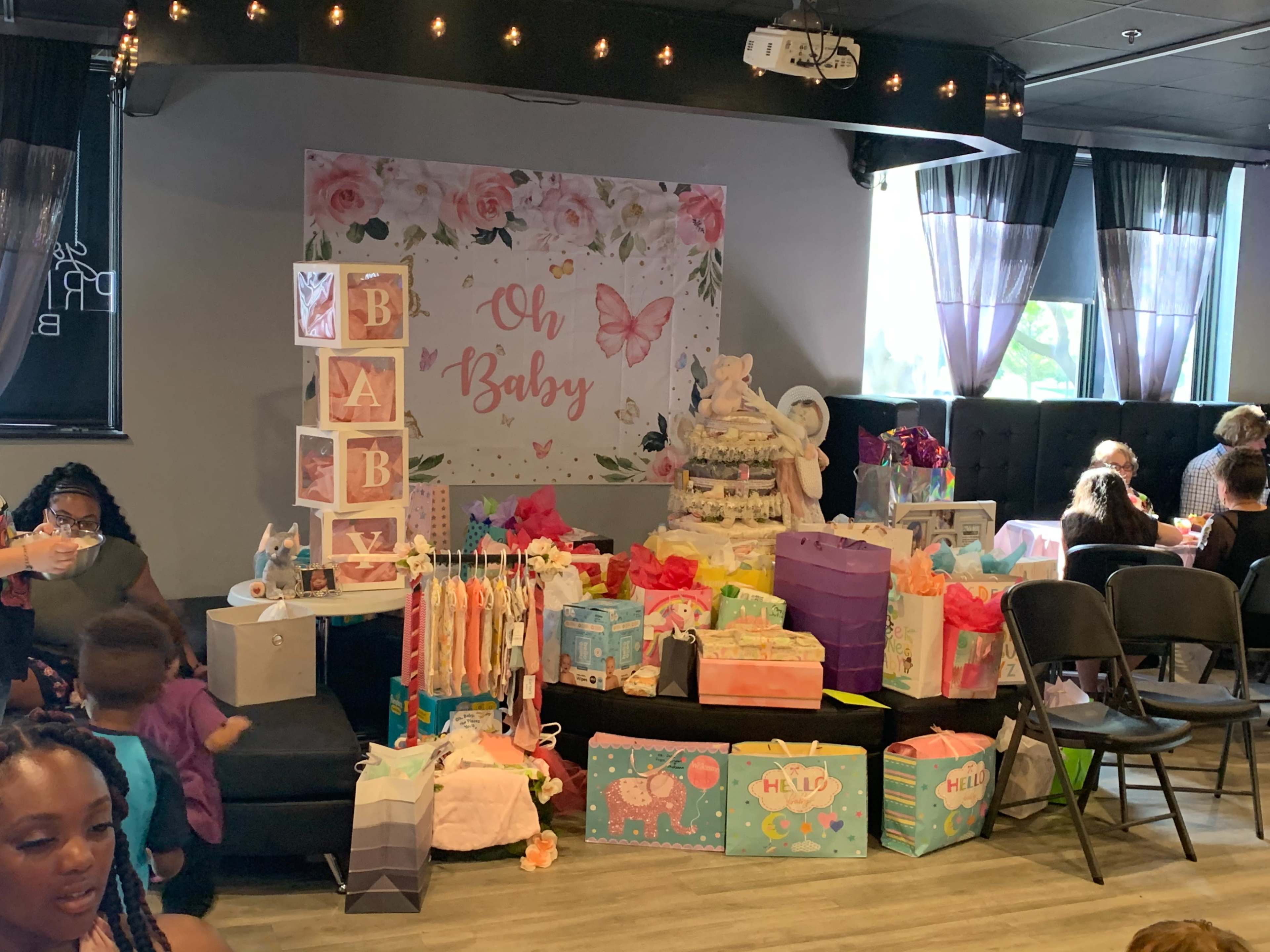 The scene shows a decorated space for a baby shower, featuring a "Oh Baby" backdrop, gift tables filled with colorful presents, and guests mingling in the background.