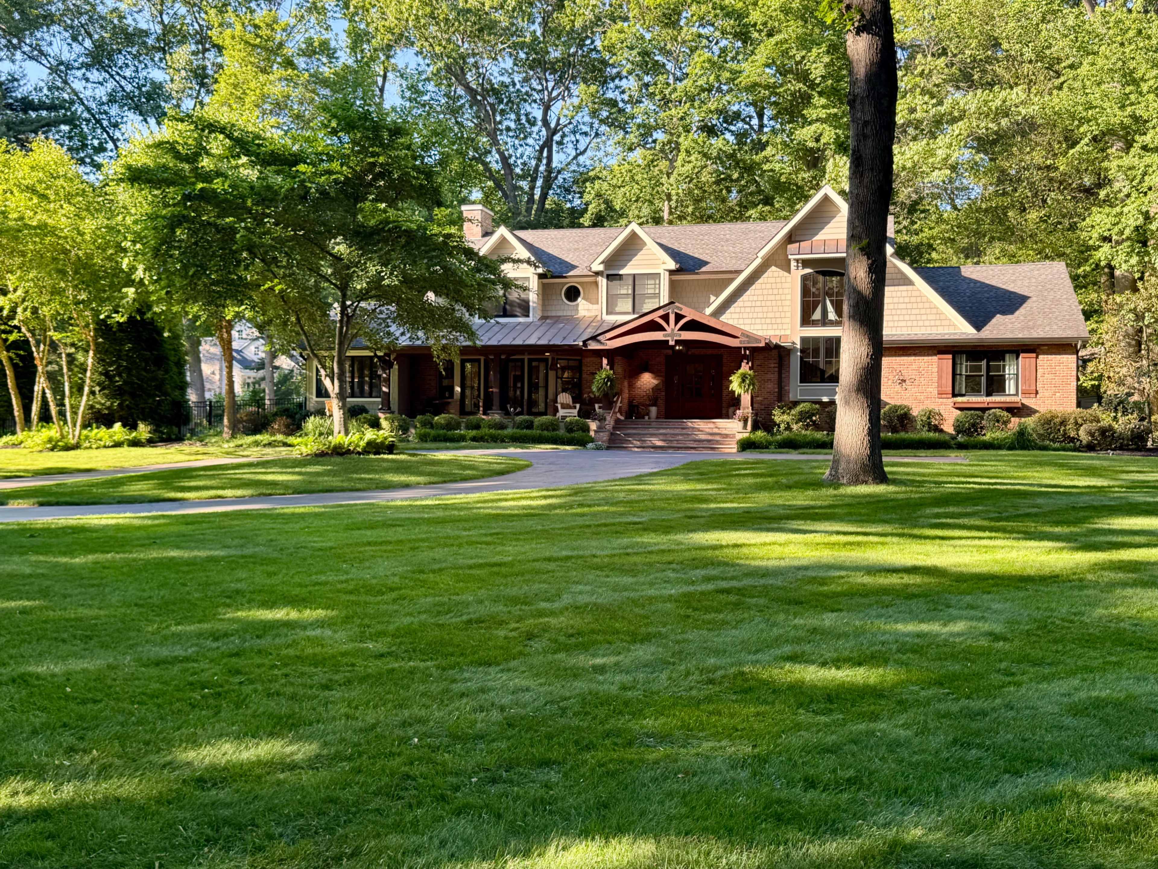 A large, two-story house with a brick exterior and a covered porch is set in a green yard surrounded by trees.