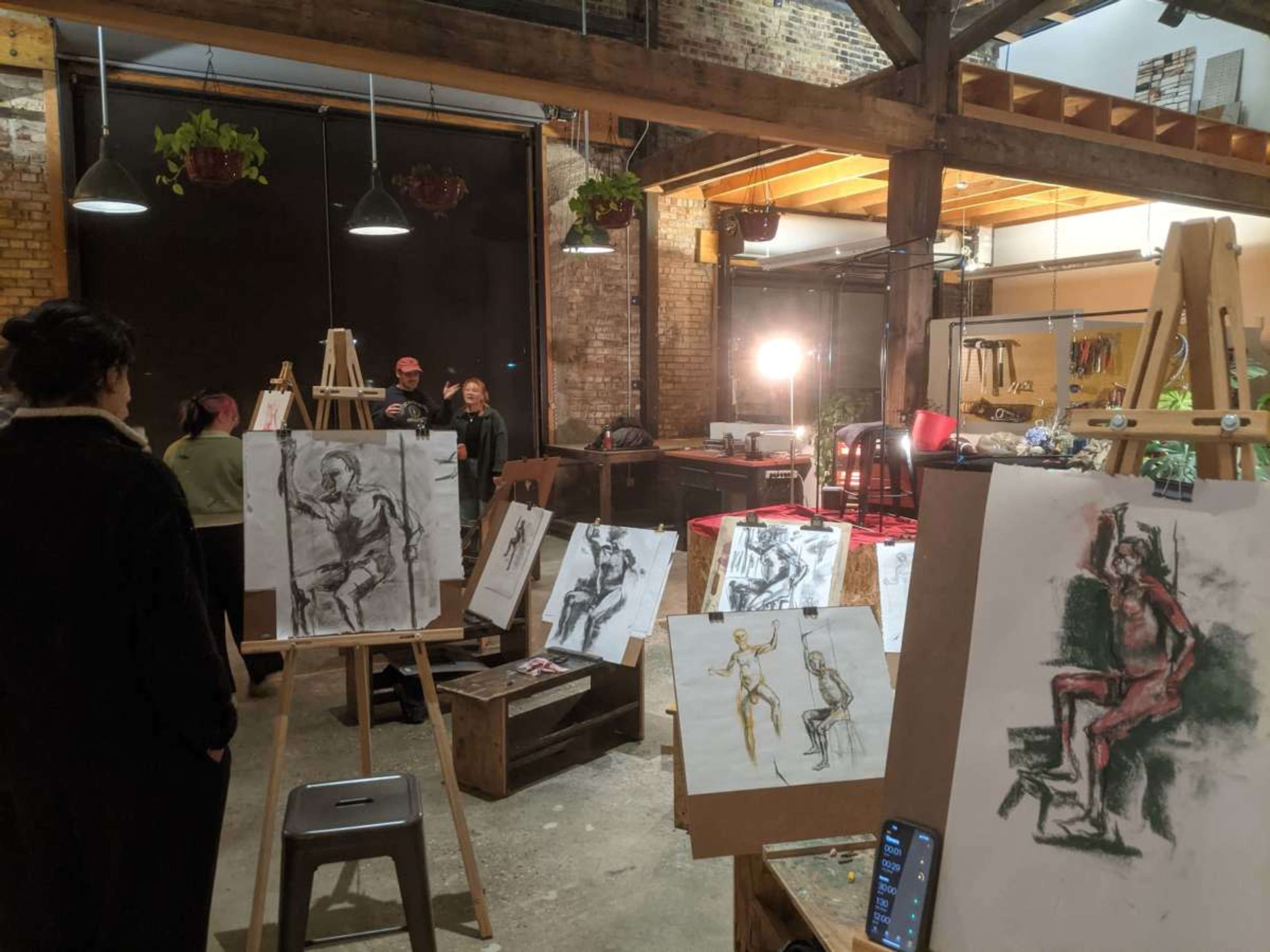 The image shows an art studio with several easels displaying sketches and charcoal drawings of human figures, while artists and attendees engage in the creative process.