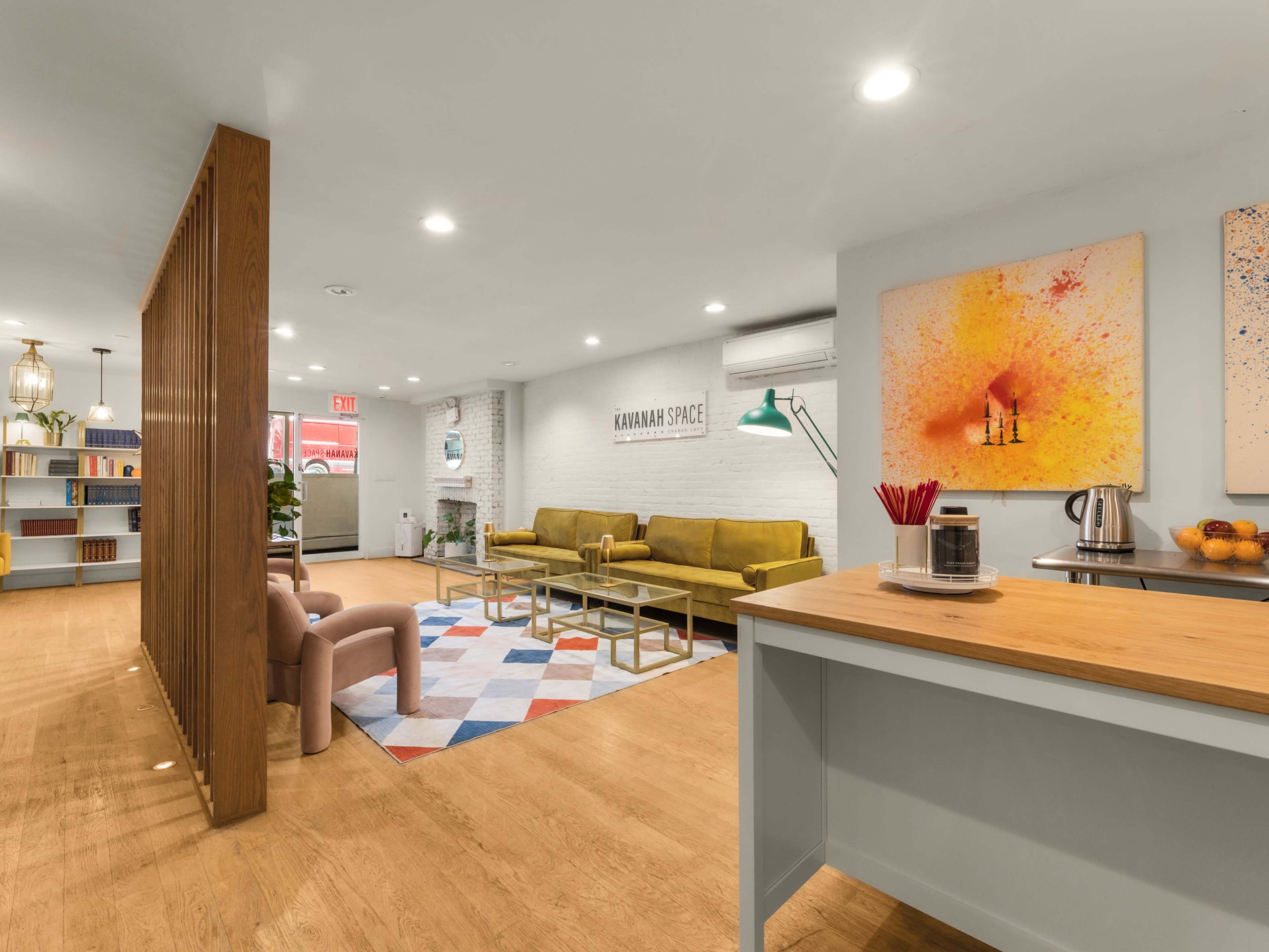 The image shows a modern communal space with seating arrangements, artwork on the walls, and a kitchenette area.