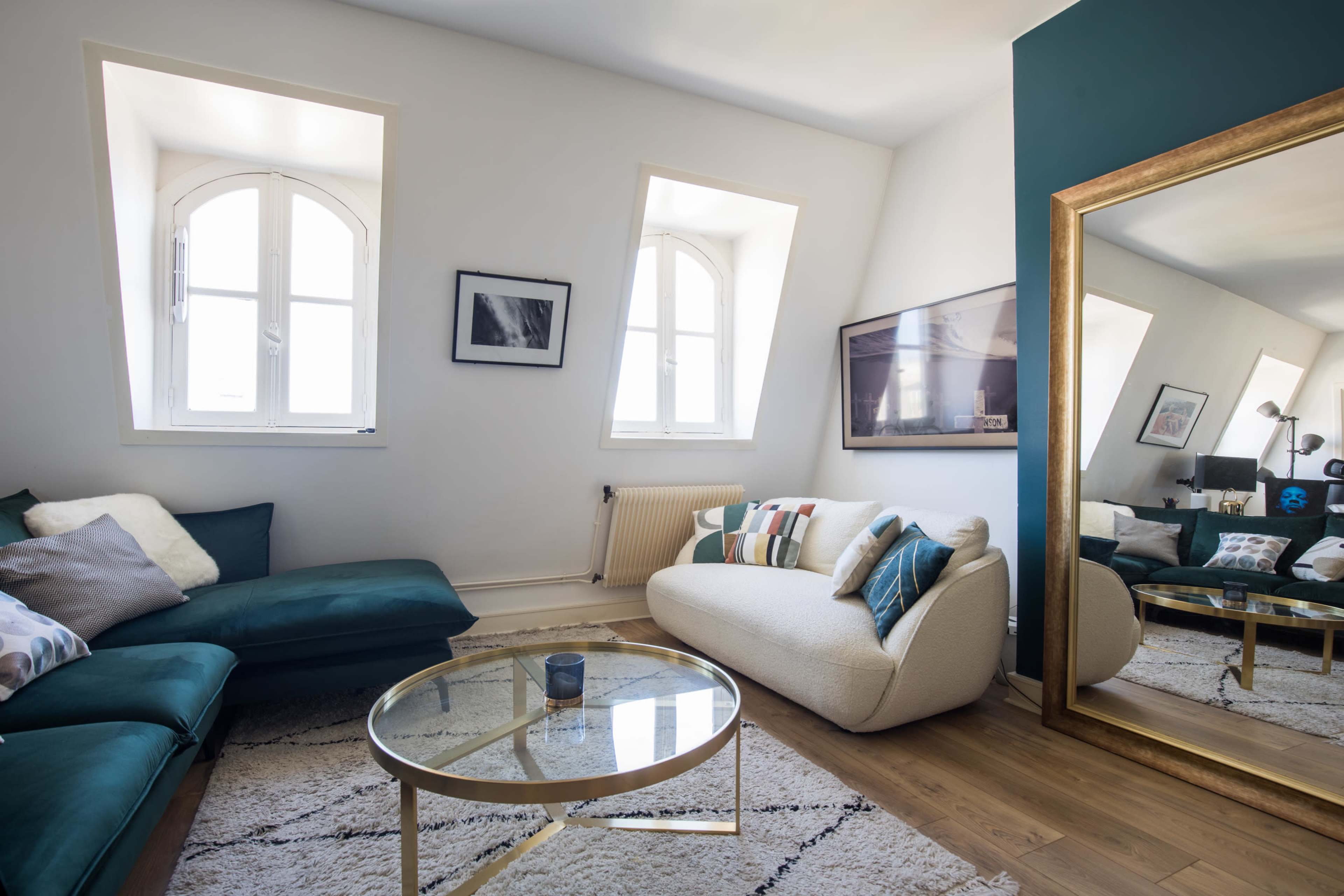 A modern living room featuring a blue sofa, a cream-colored couch, a glass coffee table, and a large mirror reflecting the space.