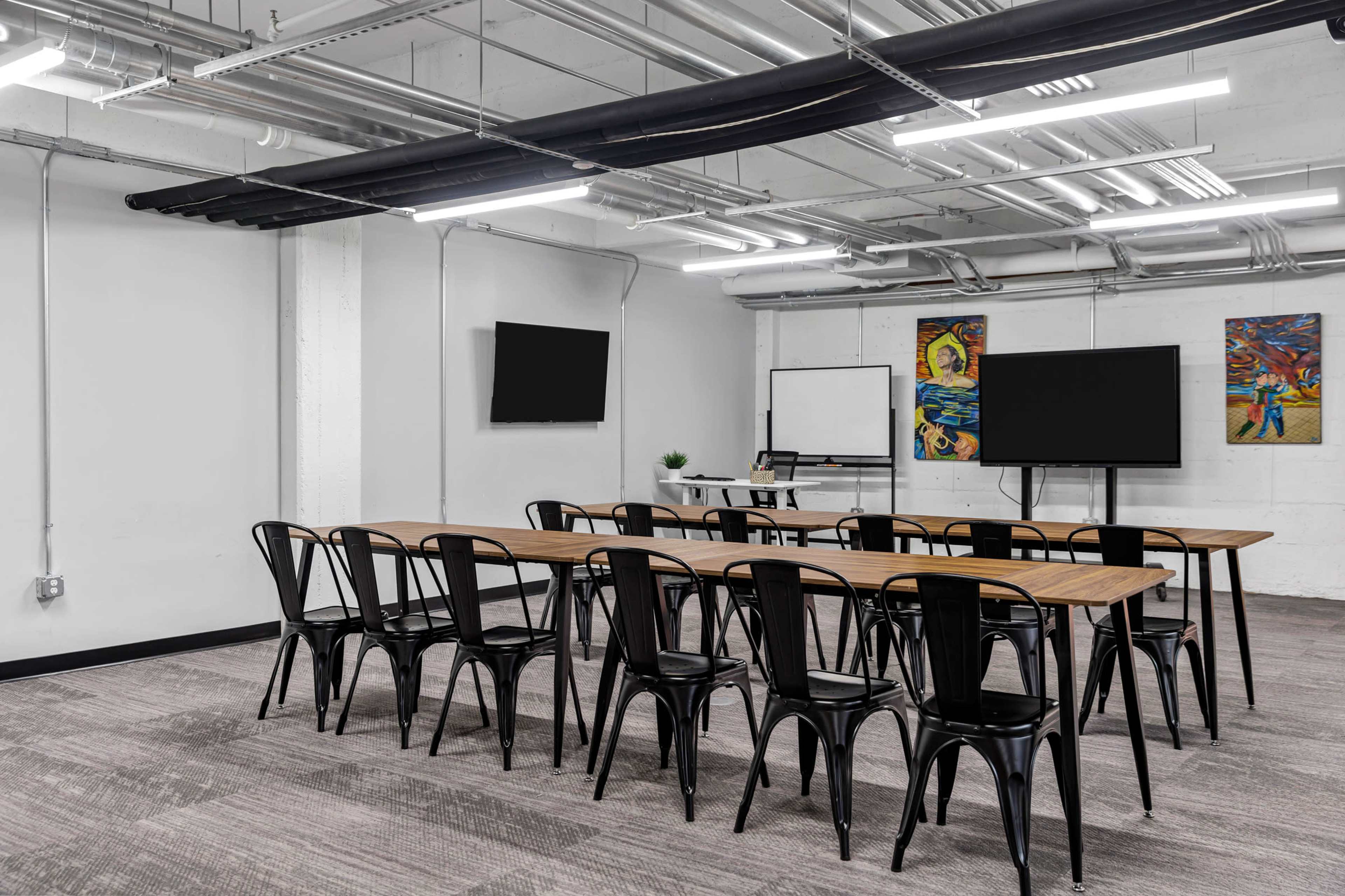 The image shows a modern conference room with two television screens, a whiteboard, and a long table surrounded by black chairs.