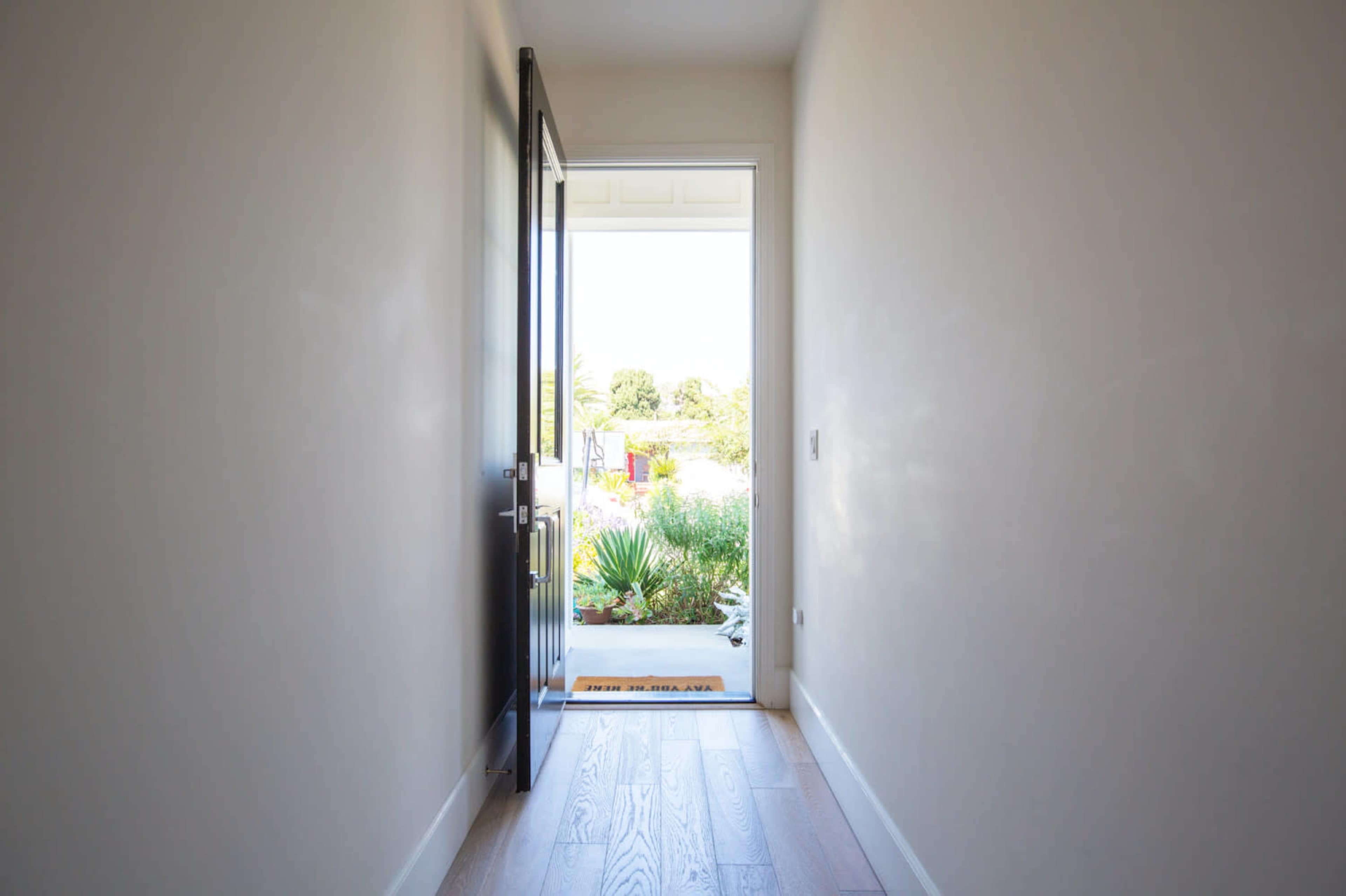 A black door is open to a well-lit hallway leading to an outdoor garden.