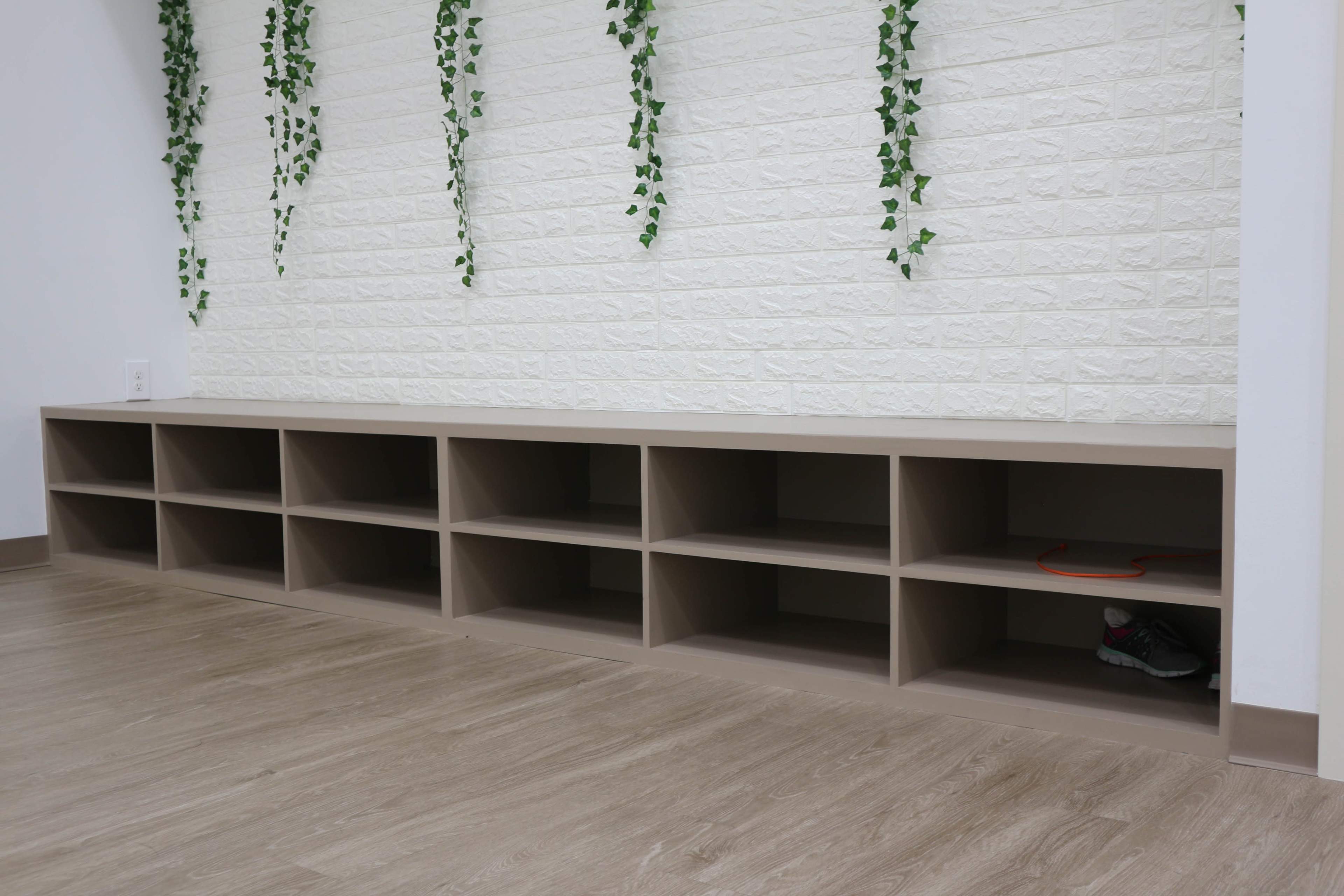 A wooden storage bench with multiple cubbies is placed against a white brick wall adorned with green hanging vines.