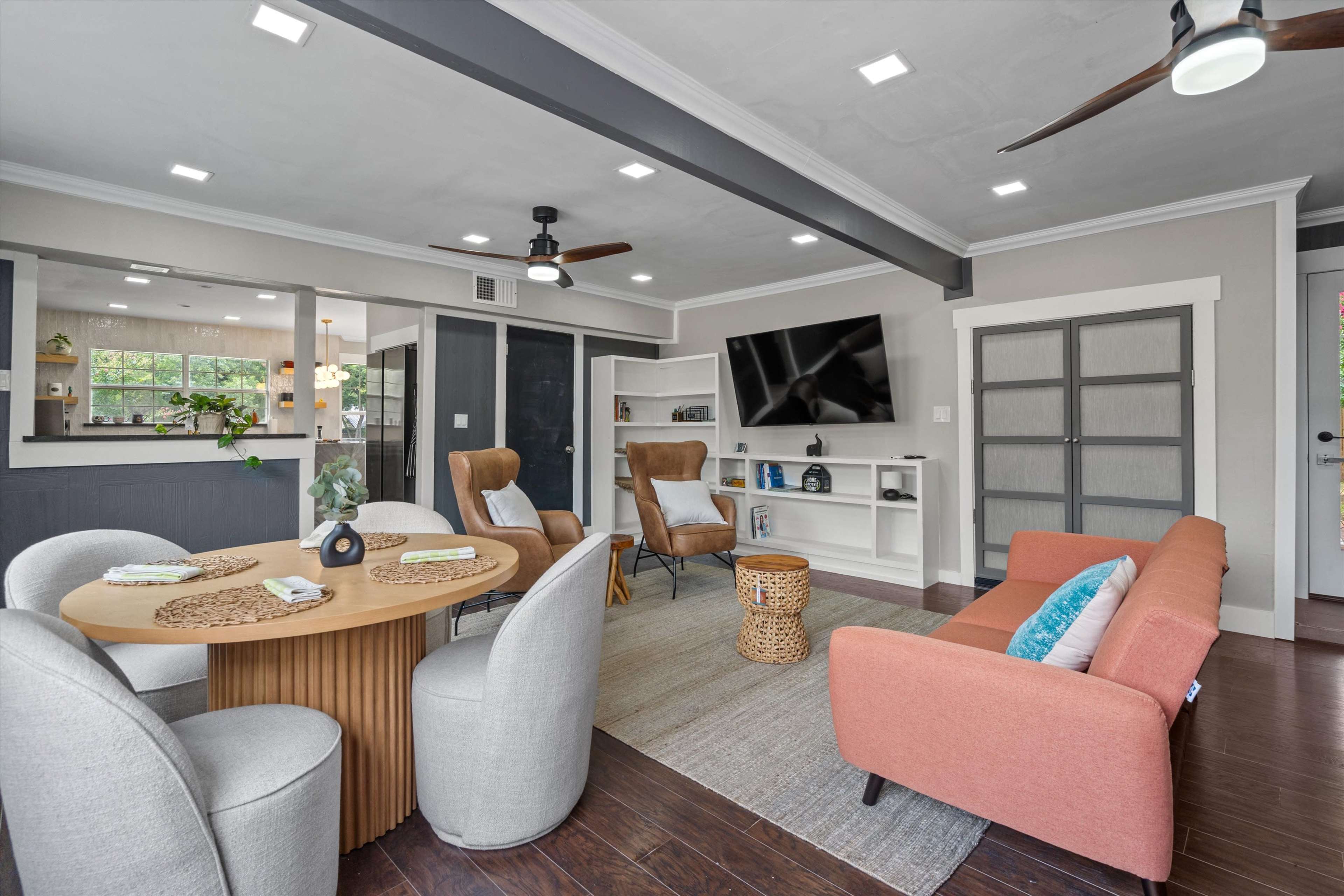 The image shows a modern living room with a circular wooden dining table, two armchairs, a sofa, and a television mounted on the wall, all arranged in a well-lit space with a neutral color scheme.