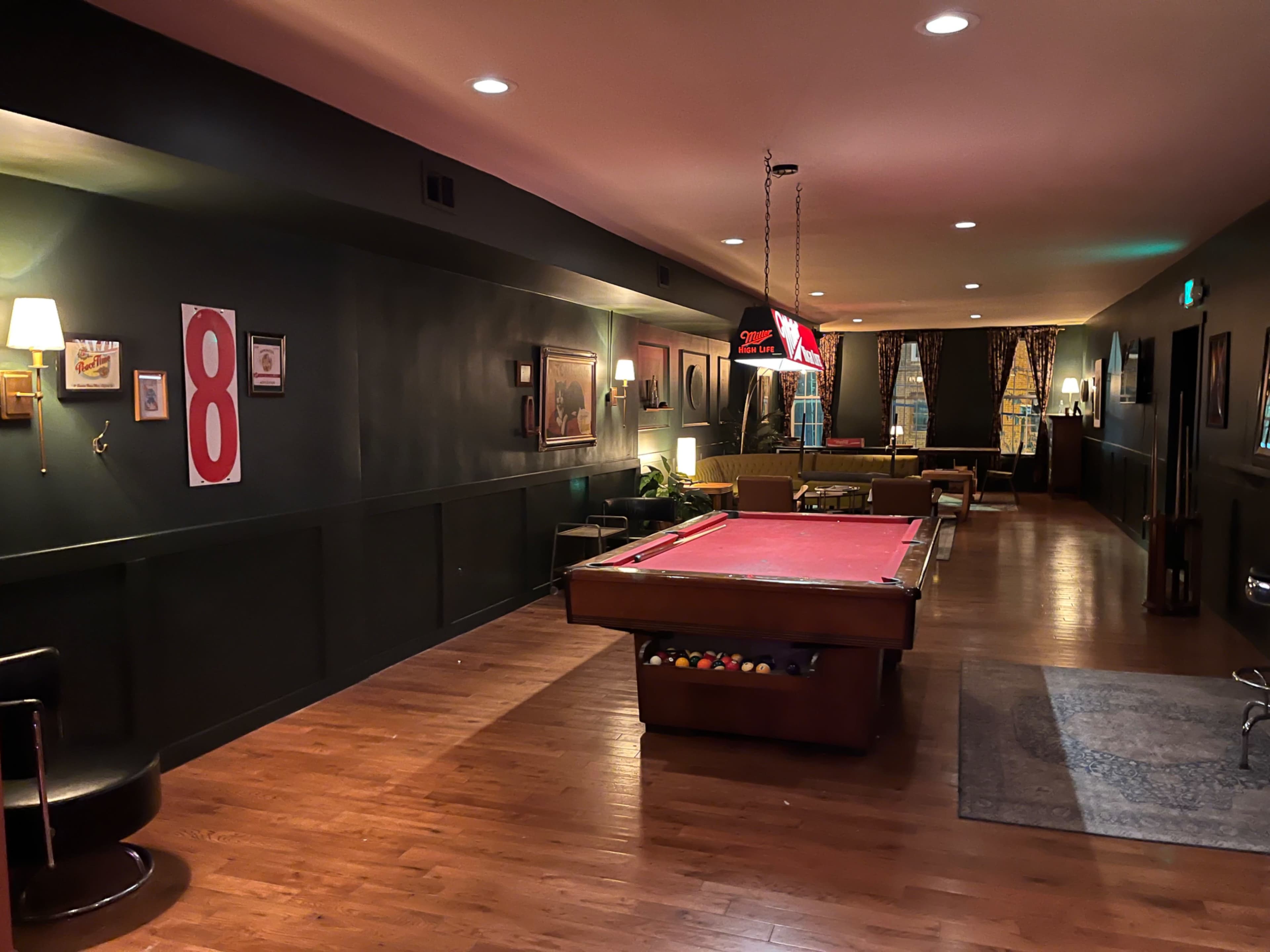 The image shows a dimly lit pool room with a red billiard table, dark walls, and a lounge area featuring two sofas and a bar.