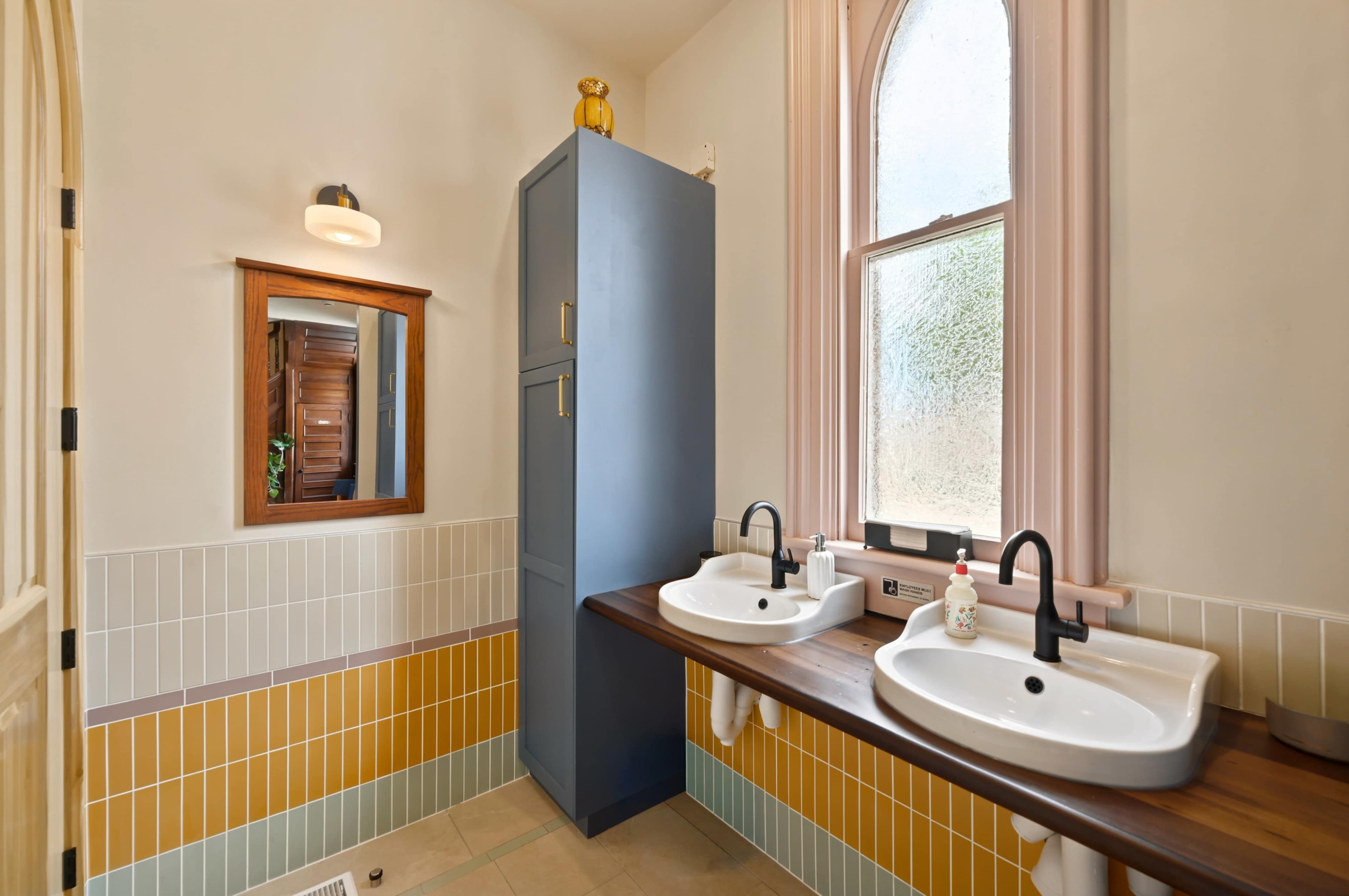 The image shows a modern bathroom with two sinks, a tall cabinet, and colorful tiled walls.