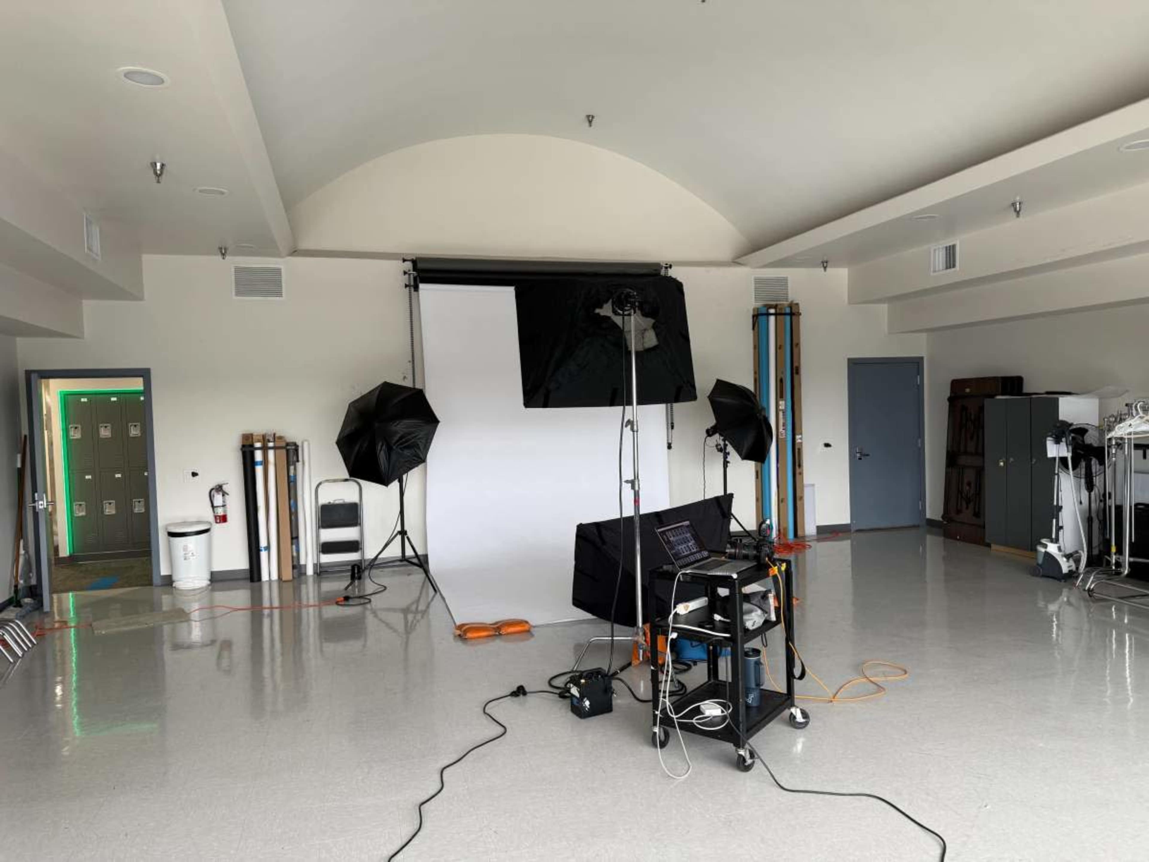 The image shows a photography setup in a spacious room, featuring a backdrop, lighting equipment, and a laptop on a cart.