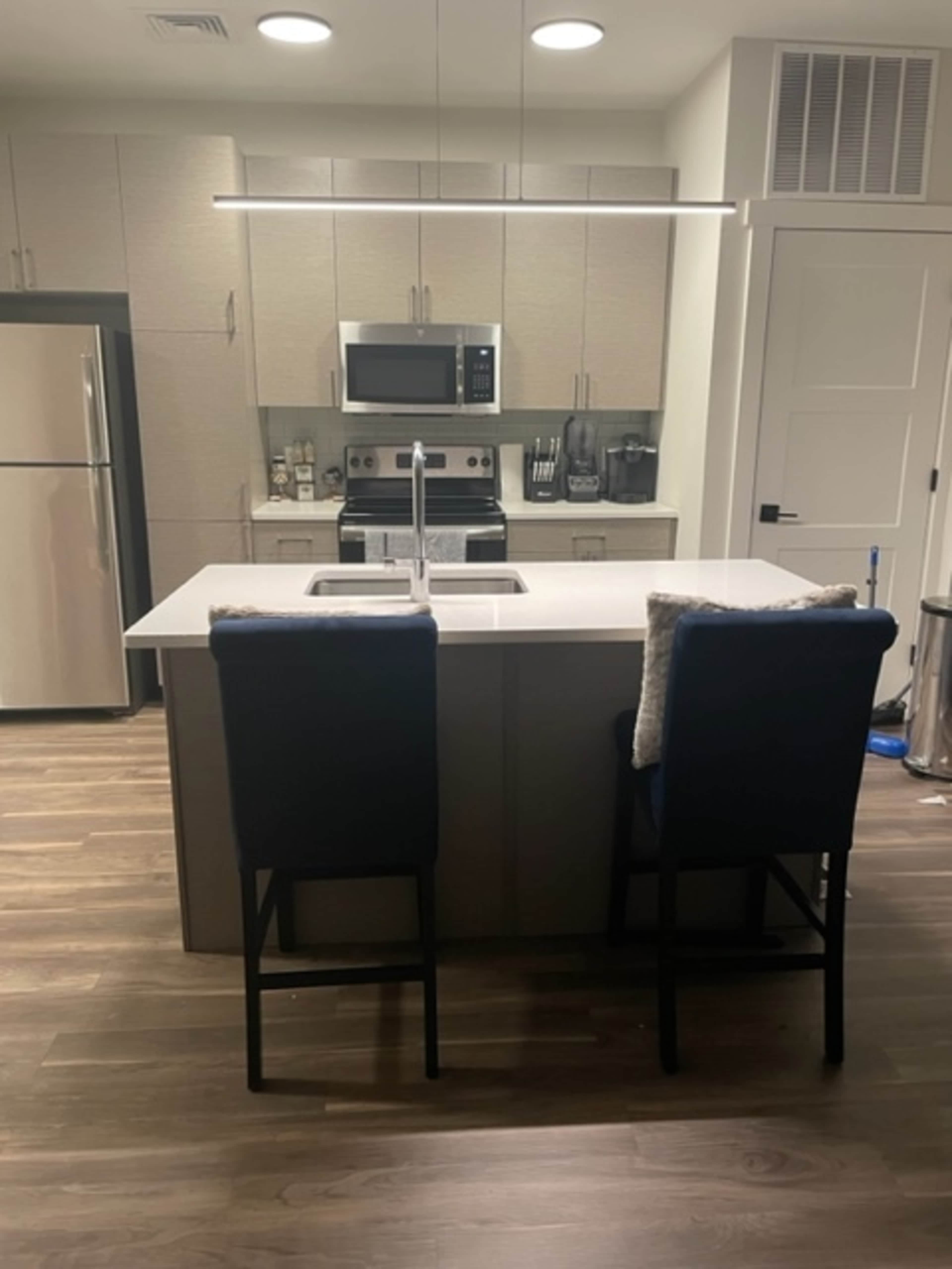 A modern kitchen with a central island featuring two bar stools, stainless steel appliances, and light wood flooring.
