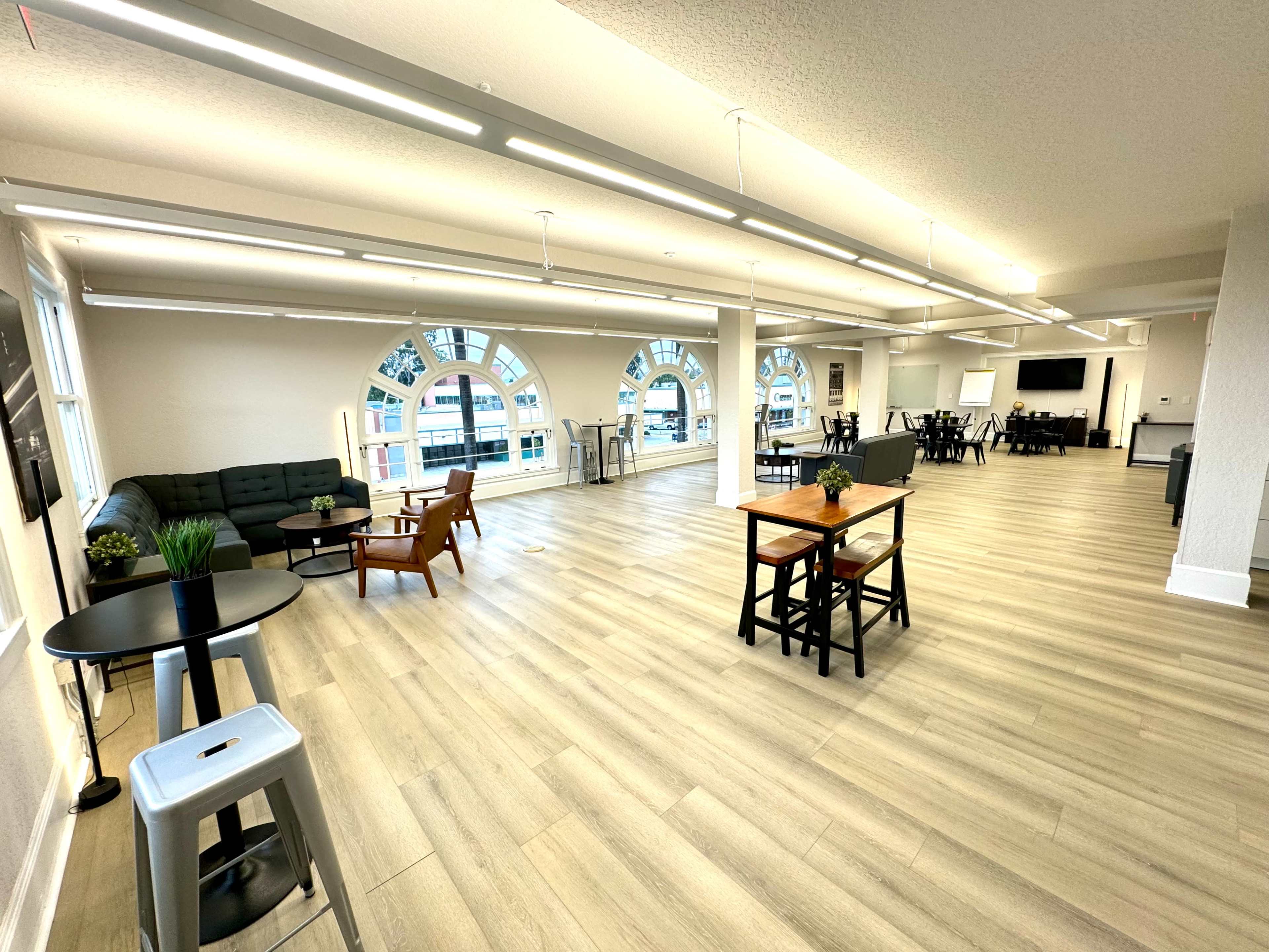 A spacious, modern interior room with multiple seating areas, large windows, and wooden flooring.