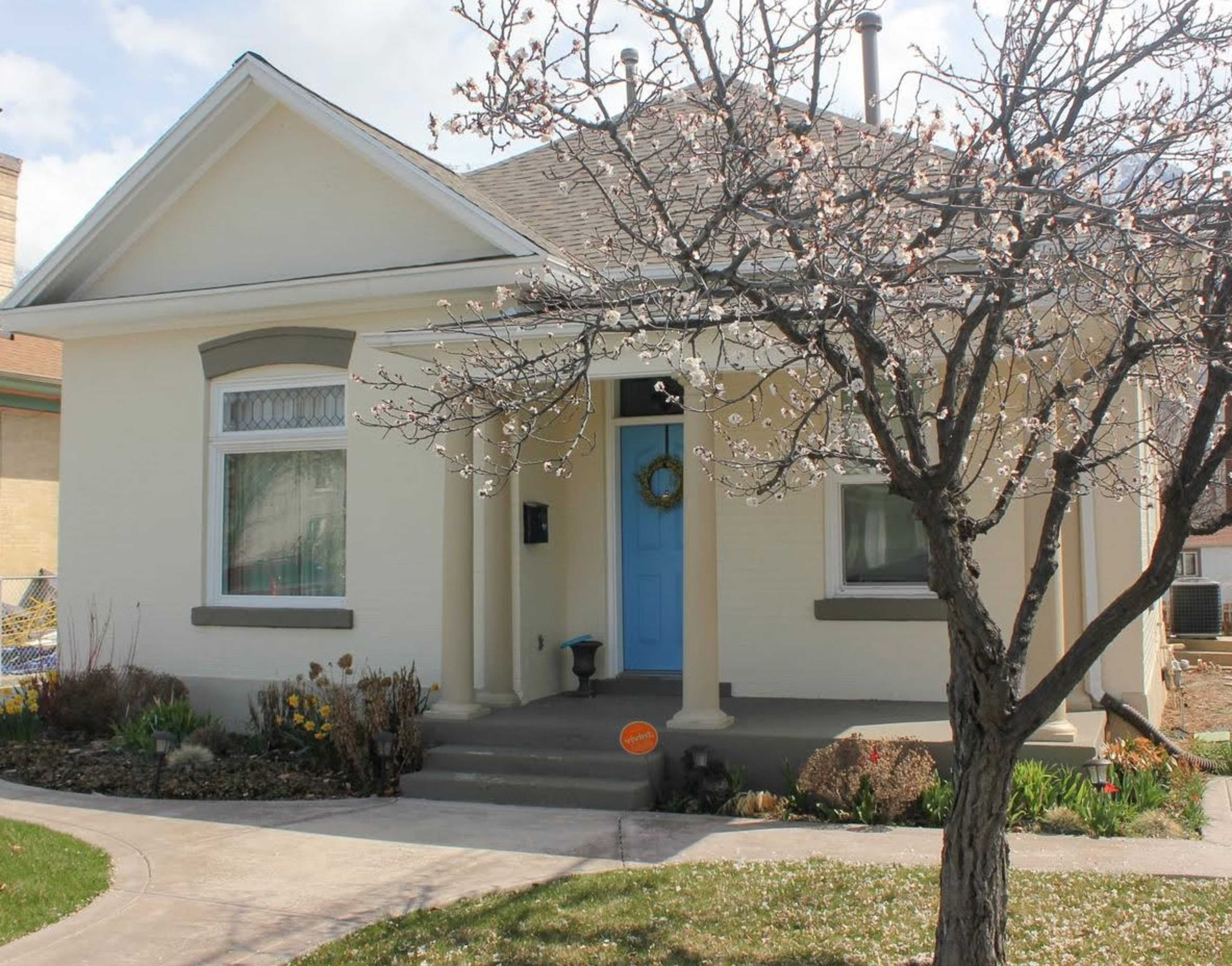 A single-story house with a front porch features a light-colored exterior, a blue door, and a flowering tree in the foreground.