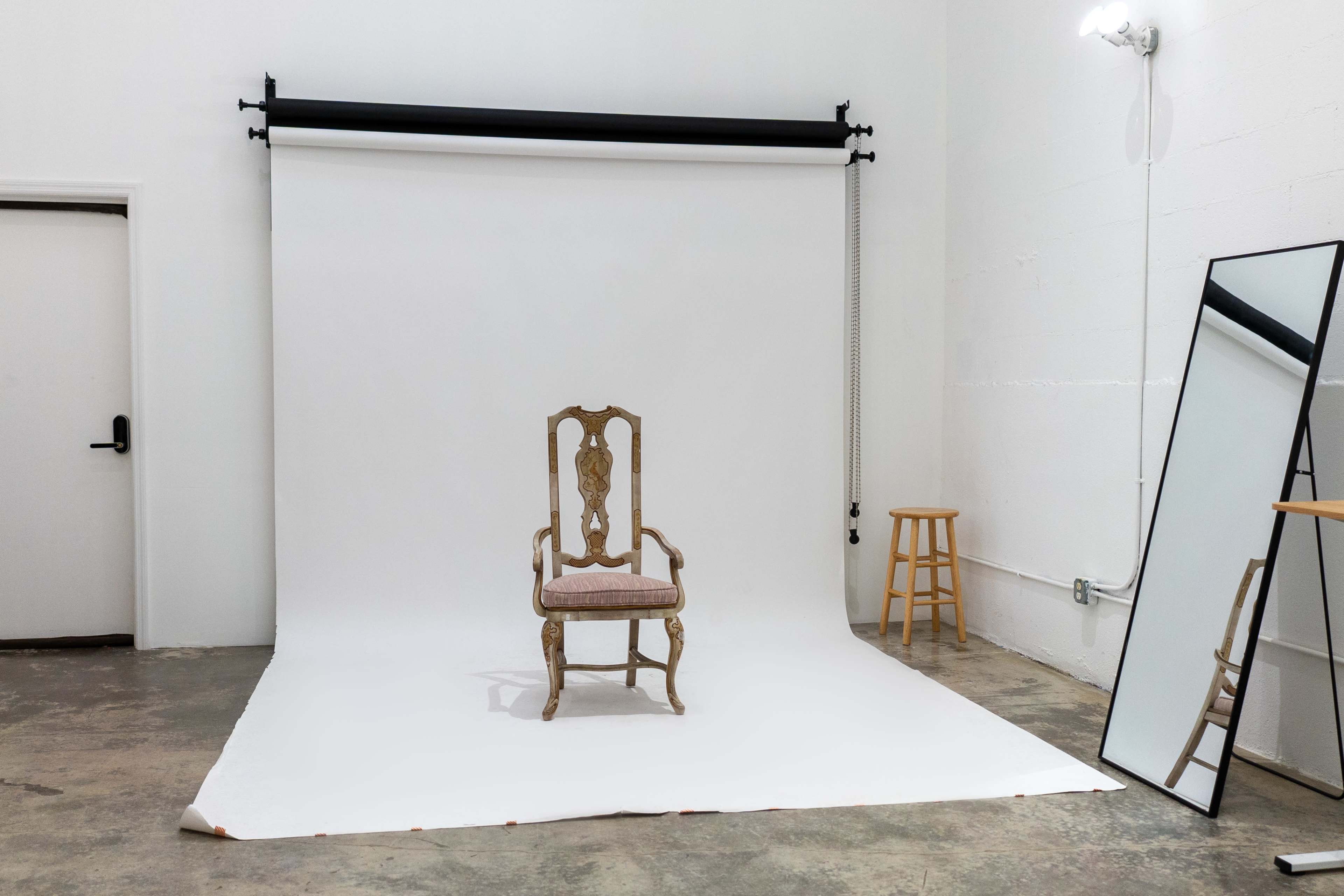 A single ornate chair is positioned in front of a white backdrop within a sparse studio space, accompanied by a large mirror and two wooden stools.