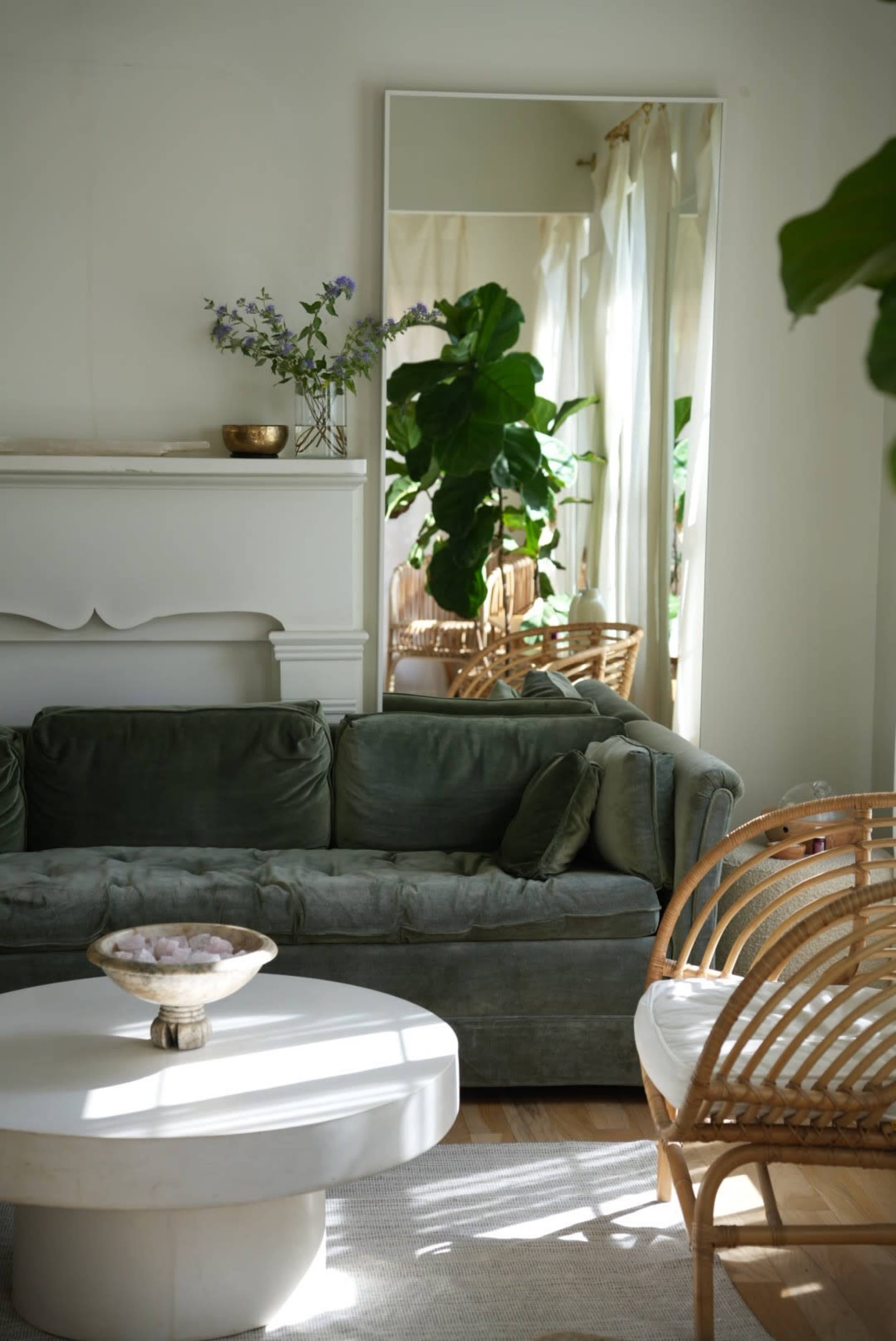 A green velvet sofa is positioned in a bright room featuring a large mirror and a round coffee table with a decorative bowl.