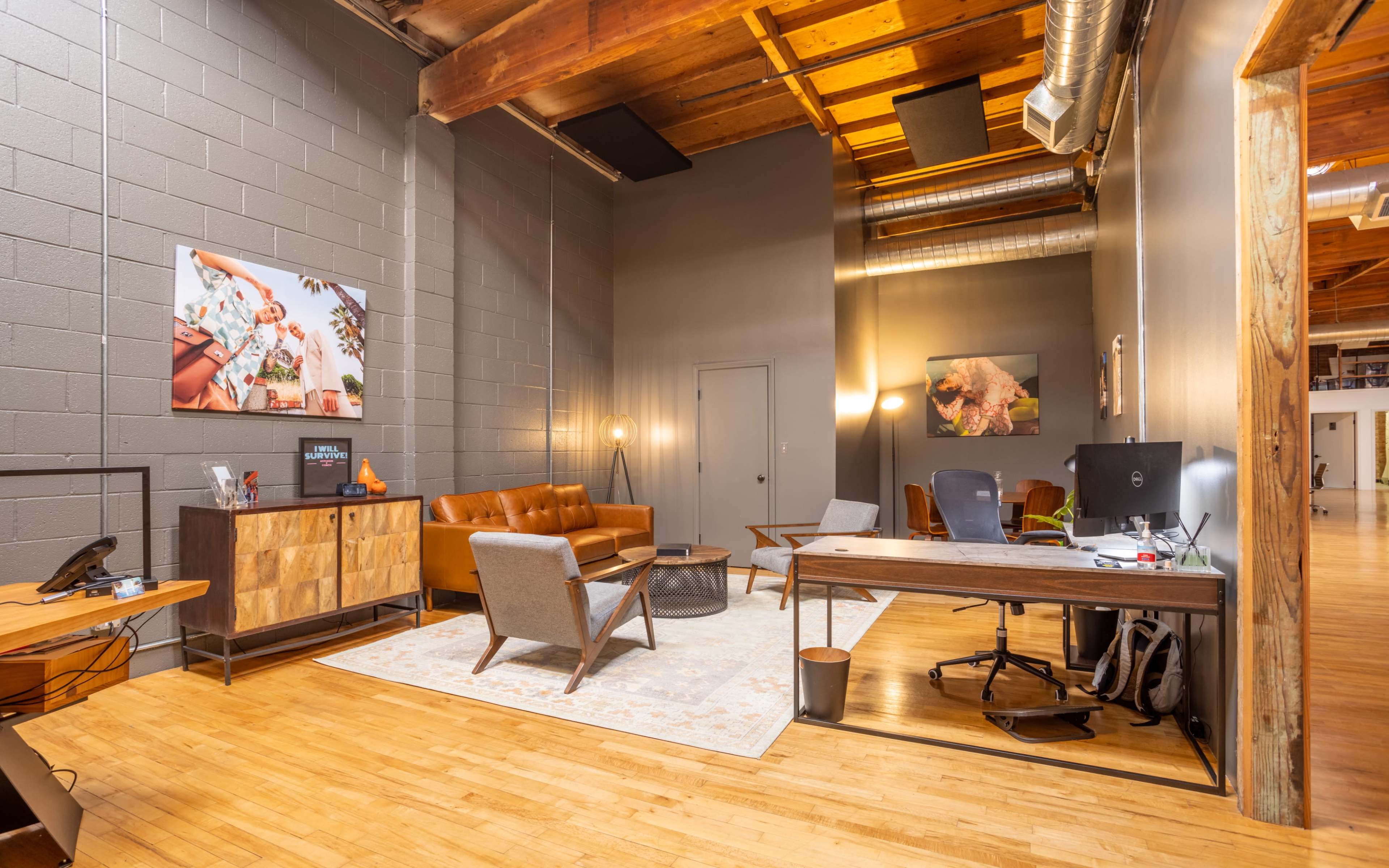 A modern office space with a brown leather couch, two chairs, a wooden desk, and a large area rug, illuminated by soft lighting.