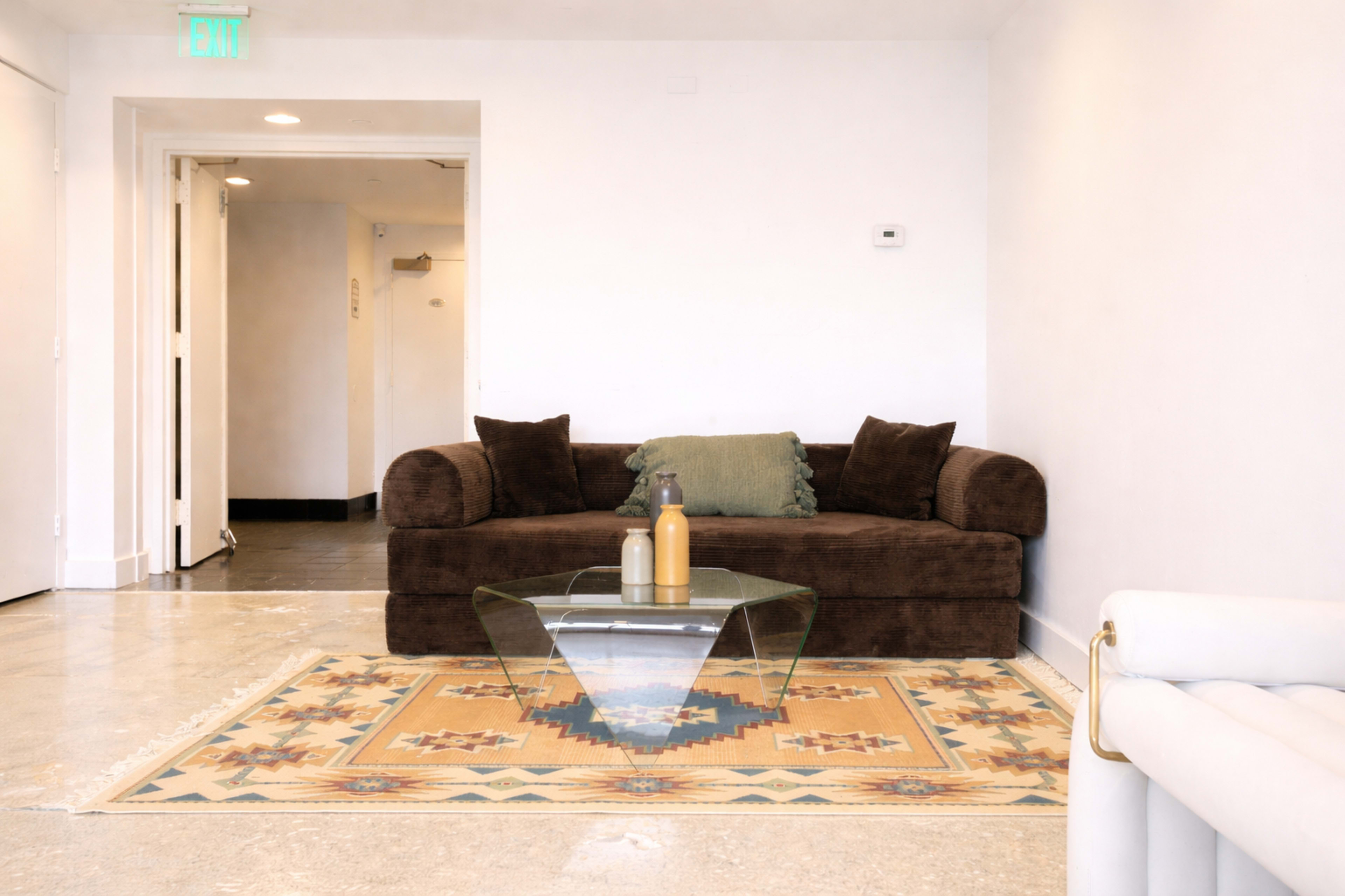 The image shows a living room with a brown sofa, a glass coffee table on a patterned rug, and a neutral wall color.