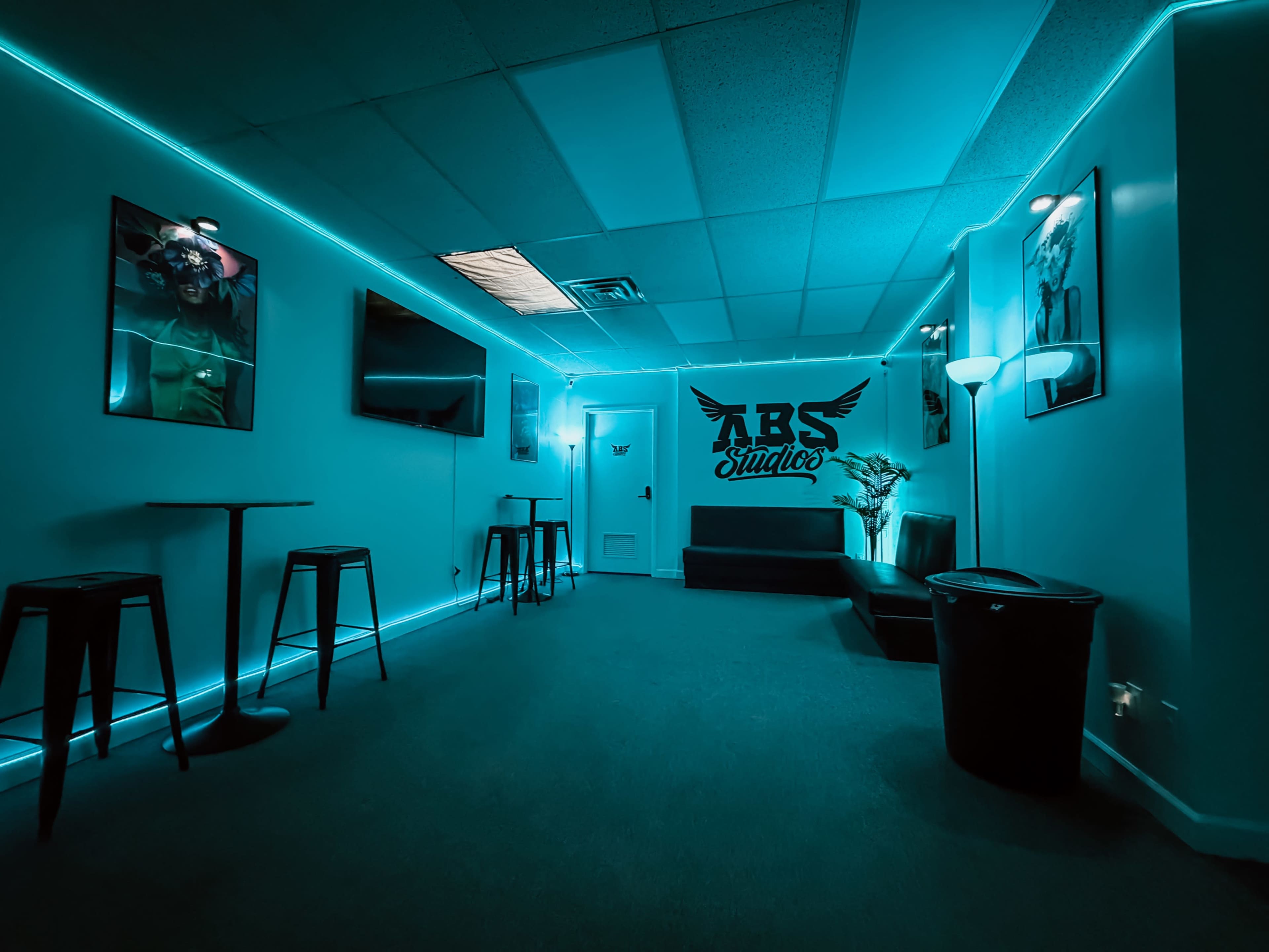 The image shows a dimly lit studio room with teal accent lighting, featuring a couch, a television, and a wall displaying the text "RBS Studio" along with artwork.