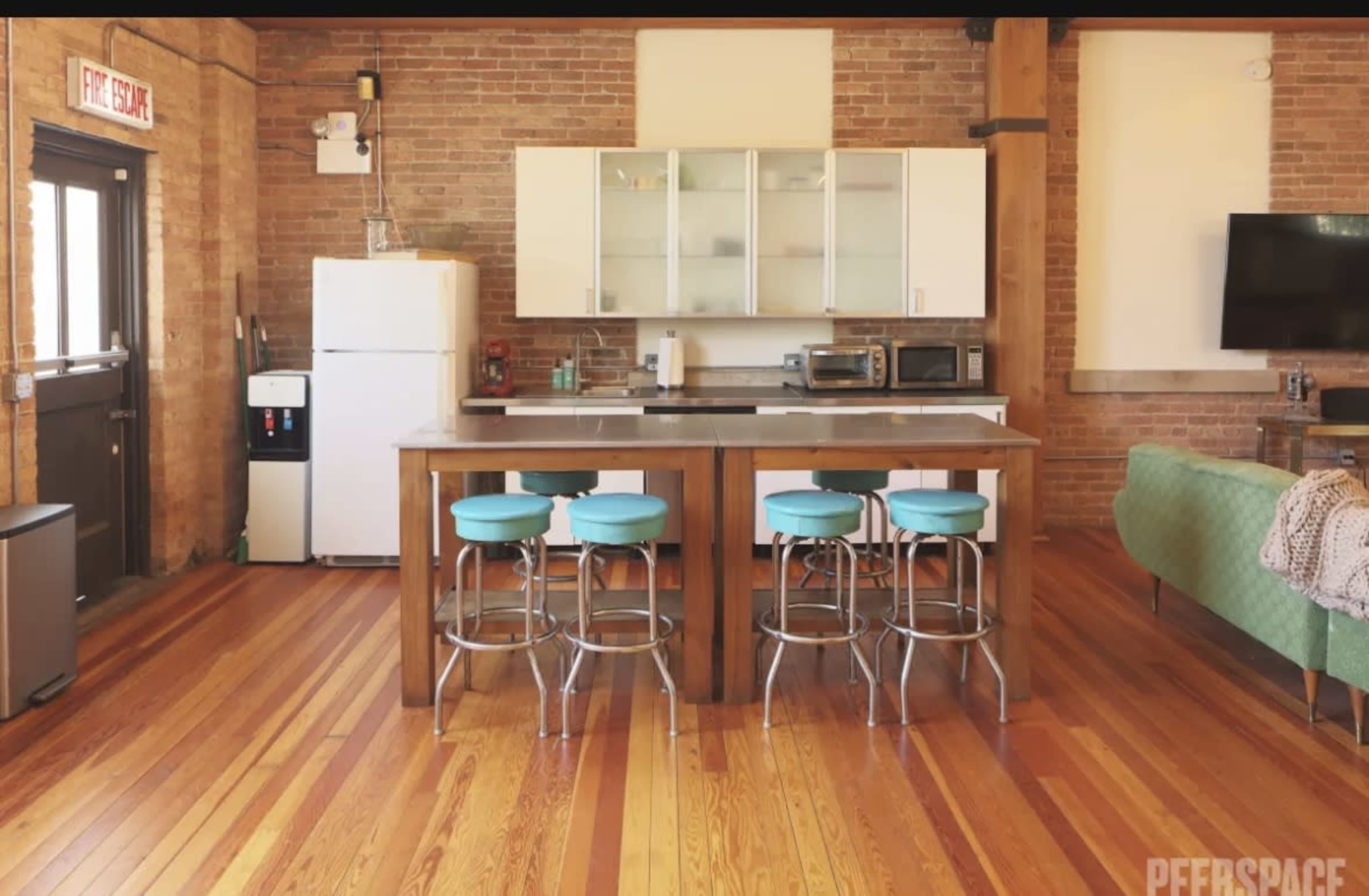 A kitchen area with a wooden island and three teal bar stools, complemented by a refrigerator, microwave, and an open living space with brick walls and hardwood floors.