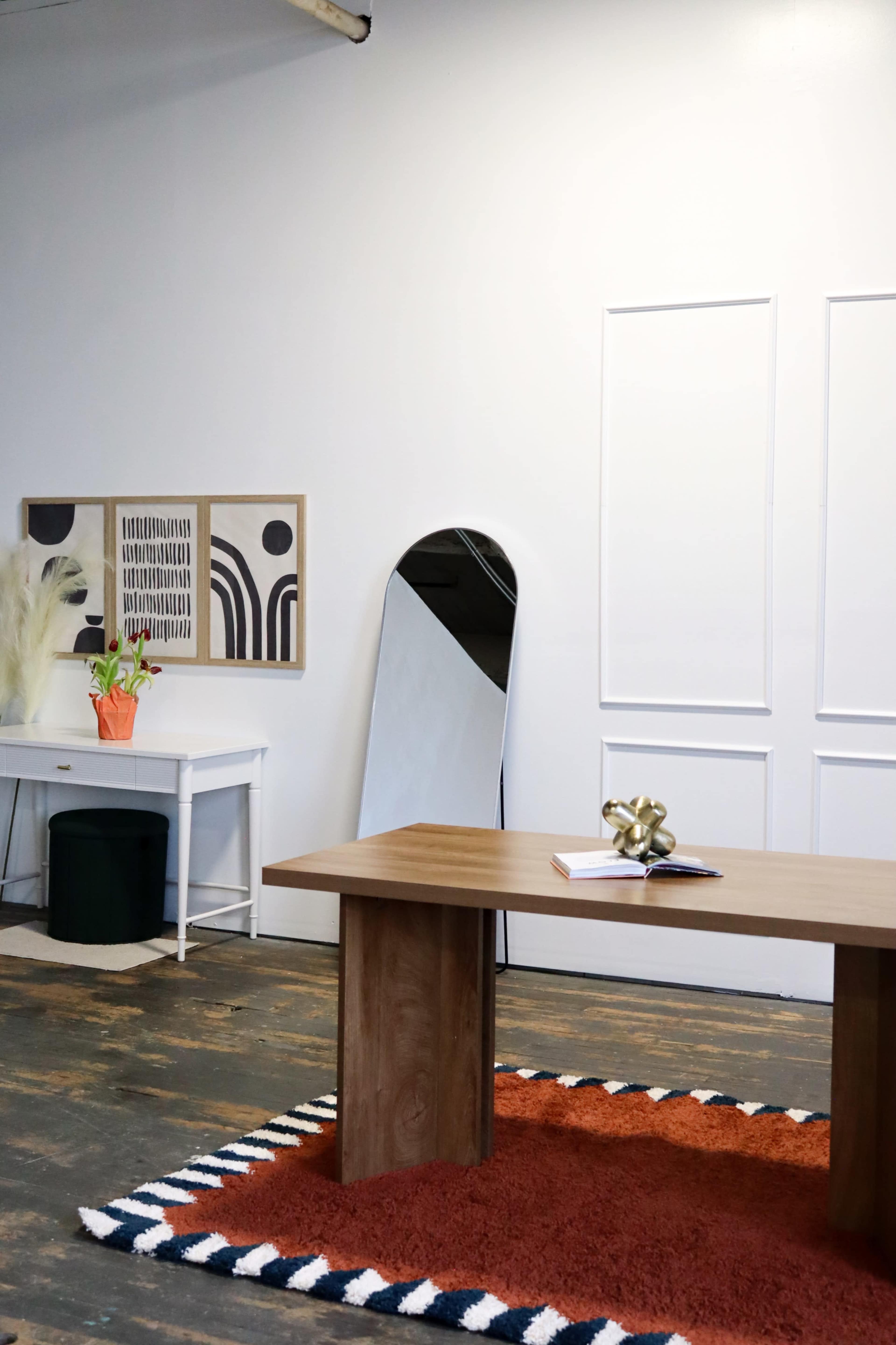 A wooden table sits on a striped rug in a minimalist room featuring a mirror, wall art, and a plant on a white side table.