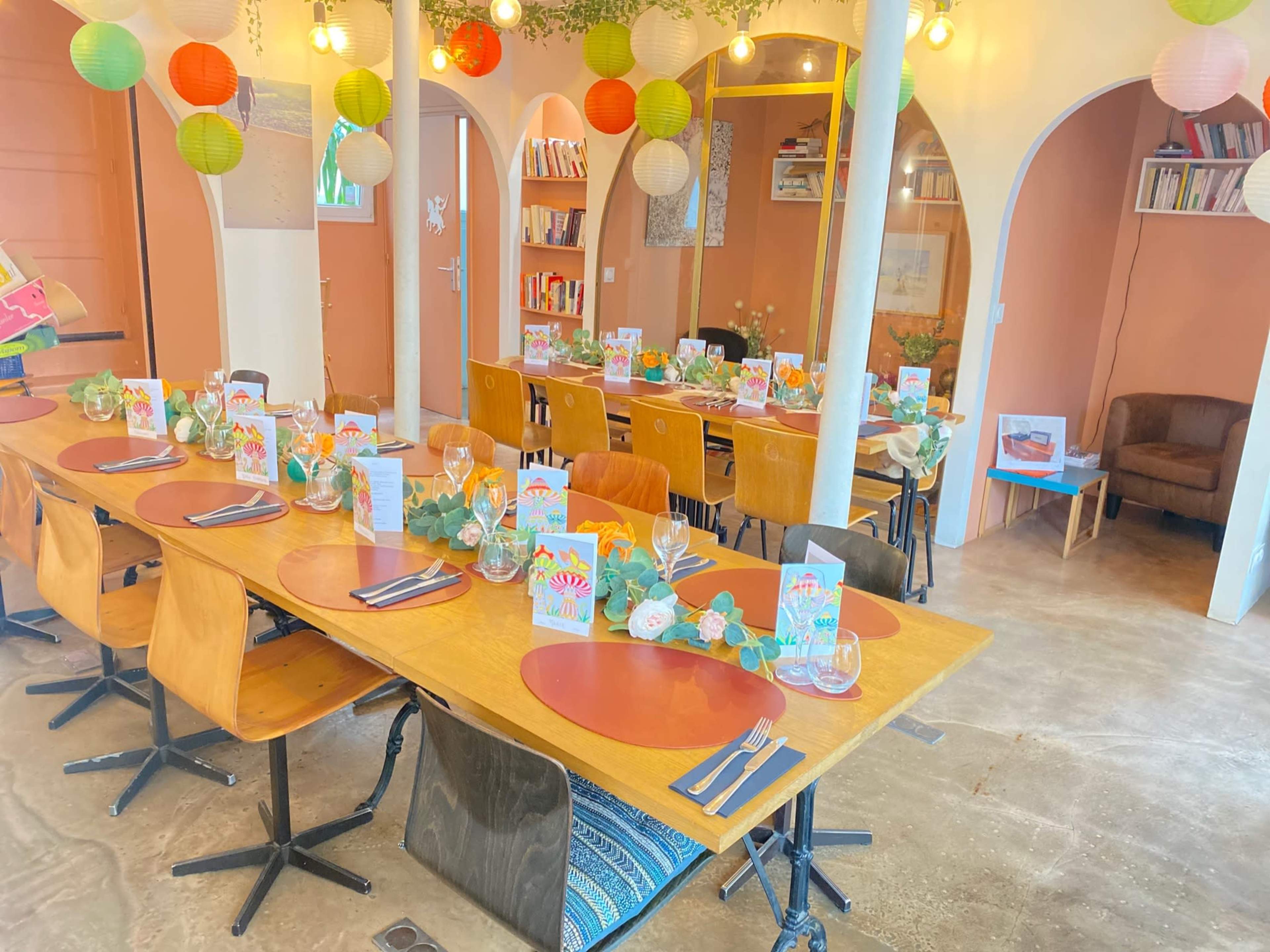 A dining area features wooden tables set with colorful menus, glassware, and decorative plants, surrounded by paper lanterns hanging from the ceiling.