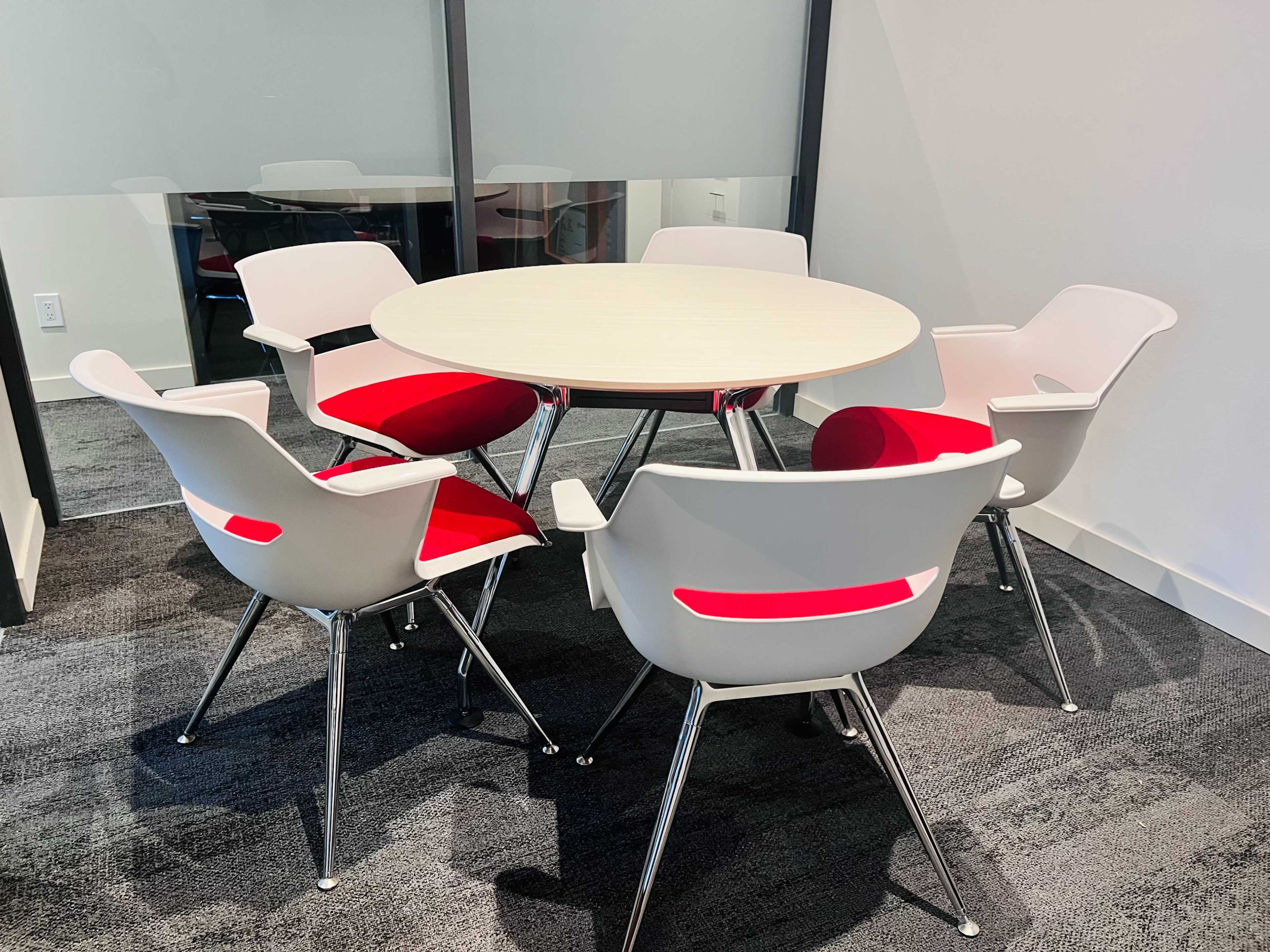 A round table surrounded by five white chairs with red seat cushions sits in a modern office setting.