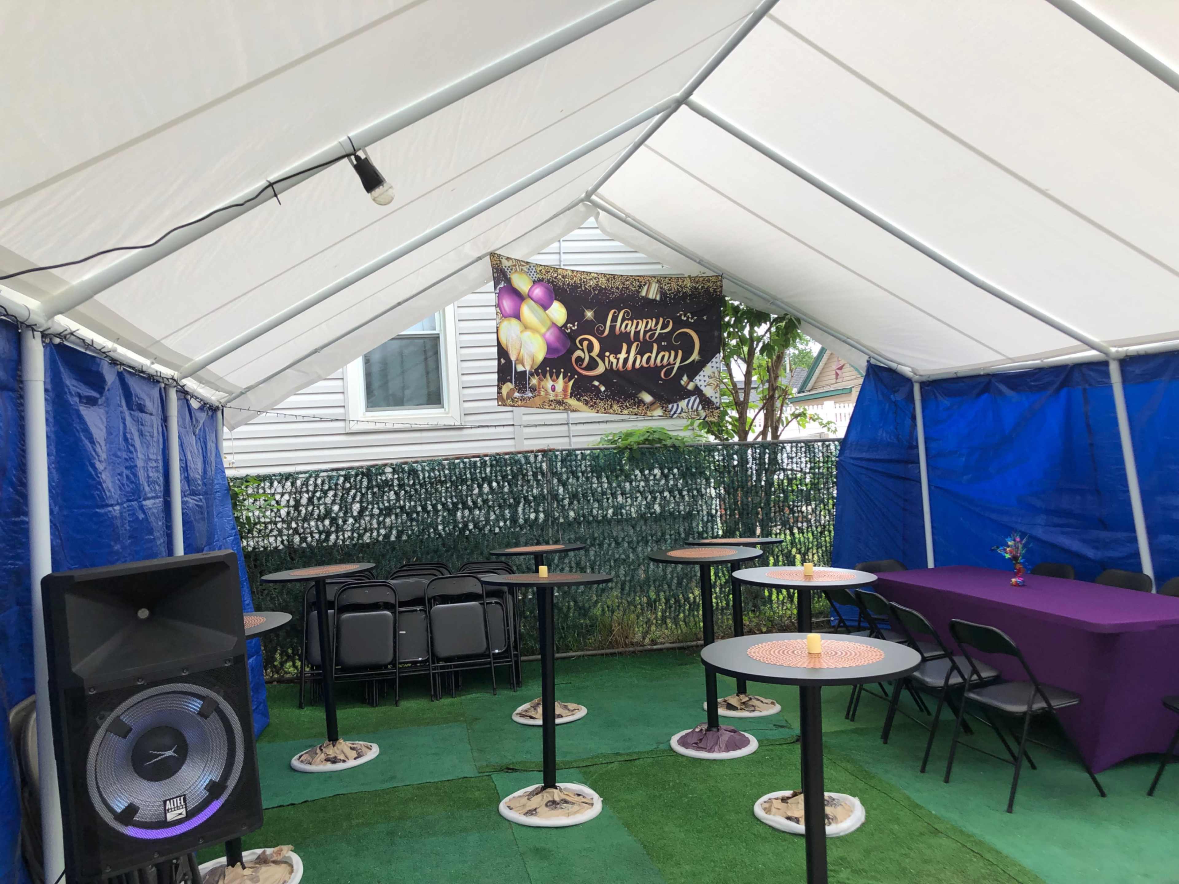 A party tent is set up with tables and chairs, featuring a "Happy Birthday" banner and a speaker in one corner.