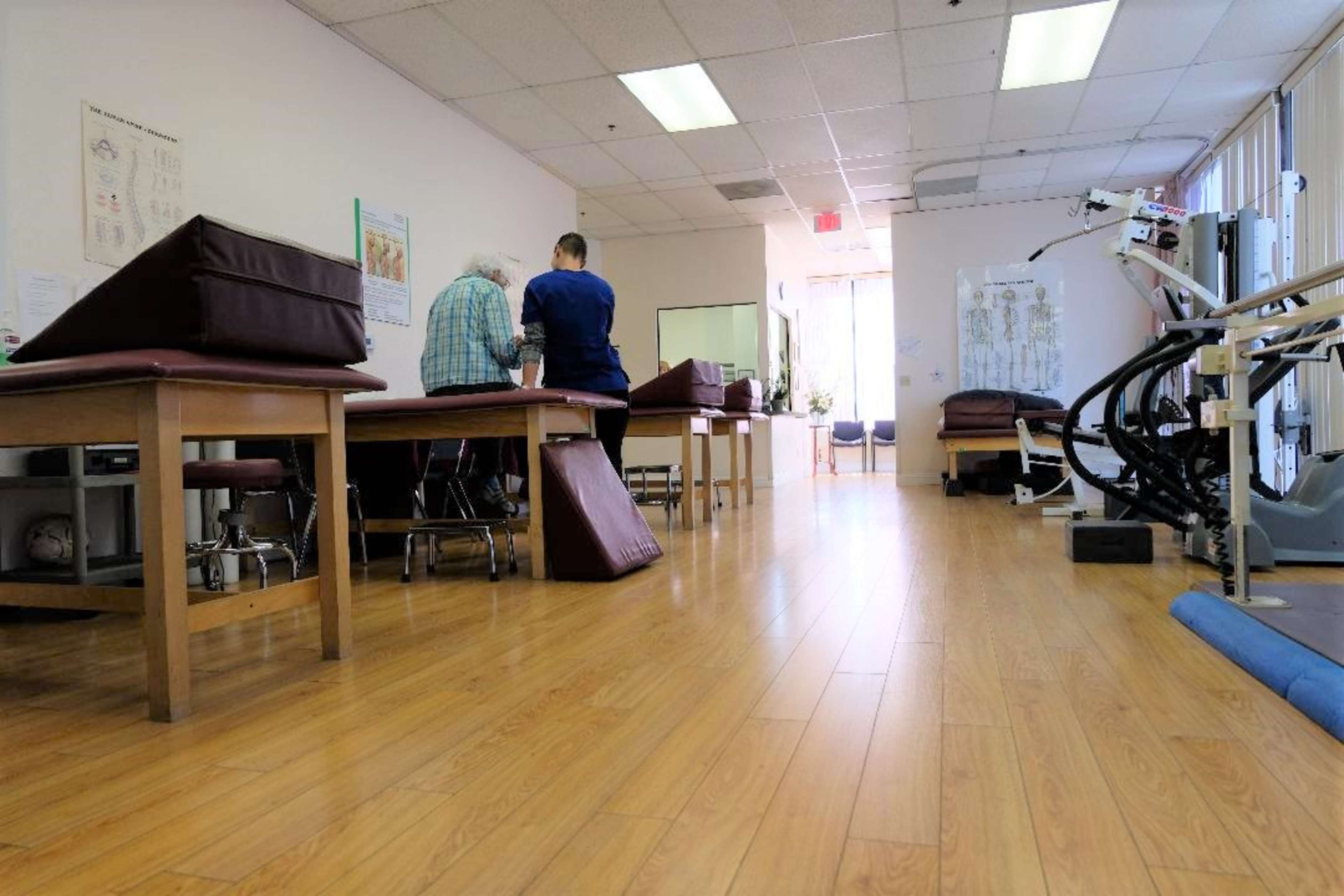 A physical therapy room features several treatment tables, a patient and therapist engaged in an activity, and equipment along the side.