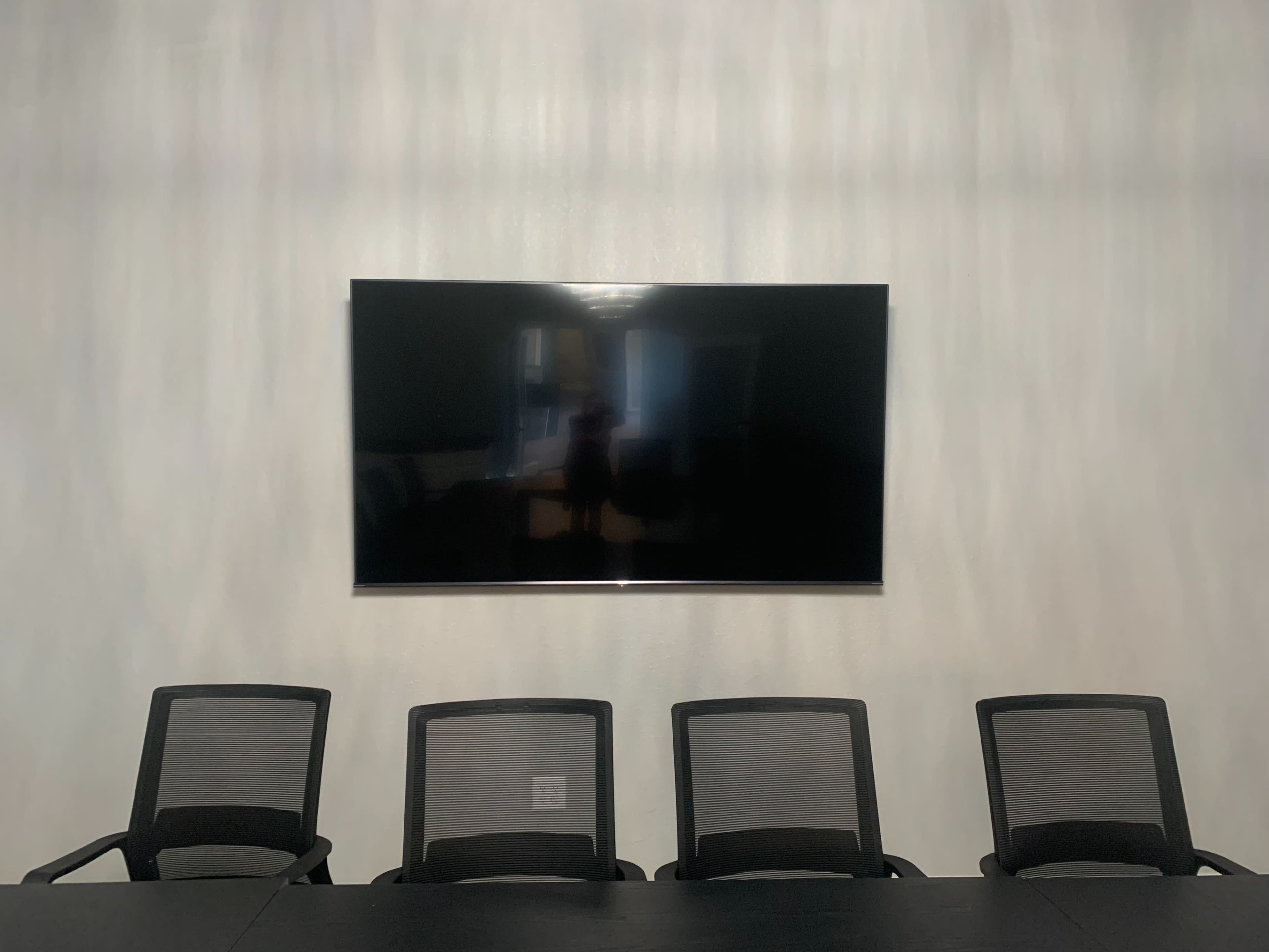 A large flat-screen television is mounted on a light-colored wall above four black mesh office chairs arranged around a dark table.